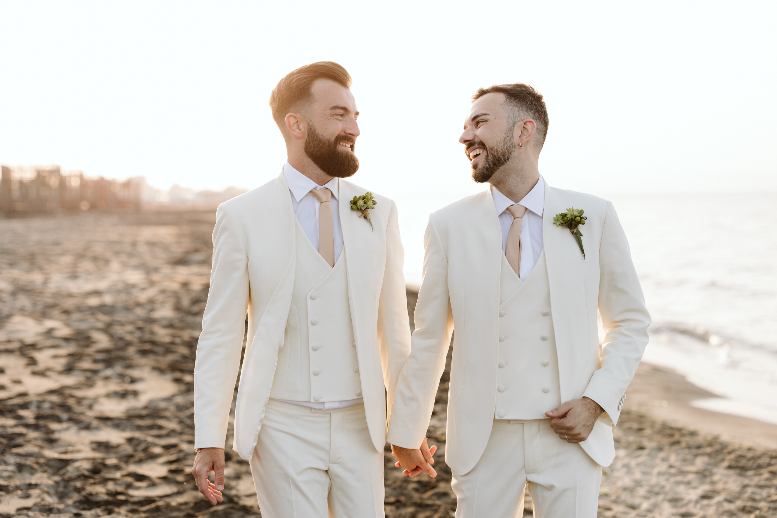 Two men in white suits holding hands and smiling at each other on a beach, with the ocean in the background.