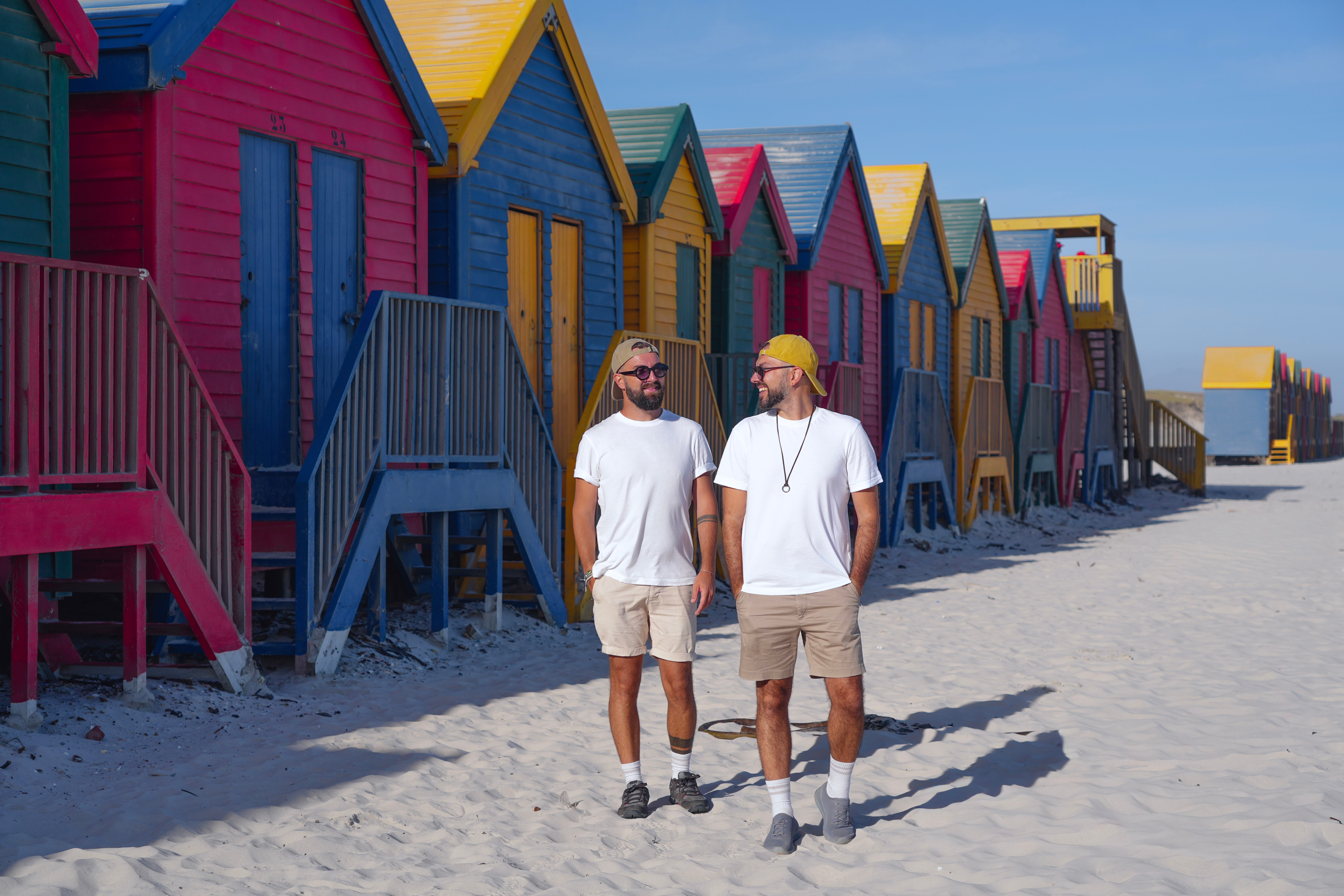 Two men walk on sandy beach near colorful beach huts, talking and smiling, with blue sky overhead.