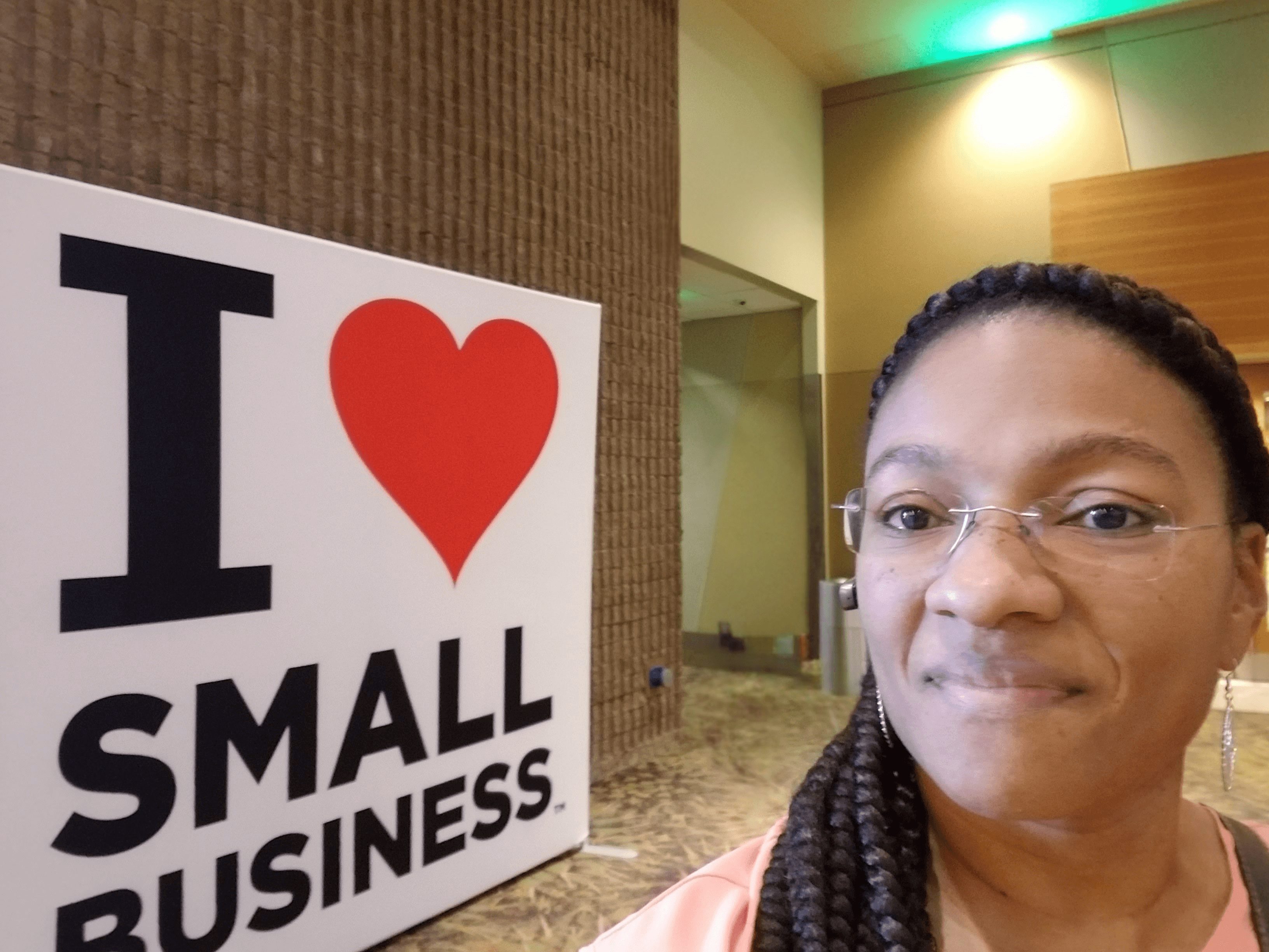 Woman with glasses and braided hair smiling indoors next to a sign that says 