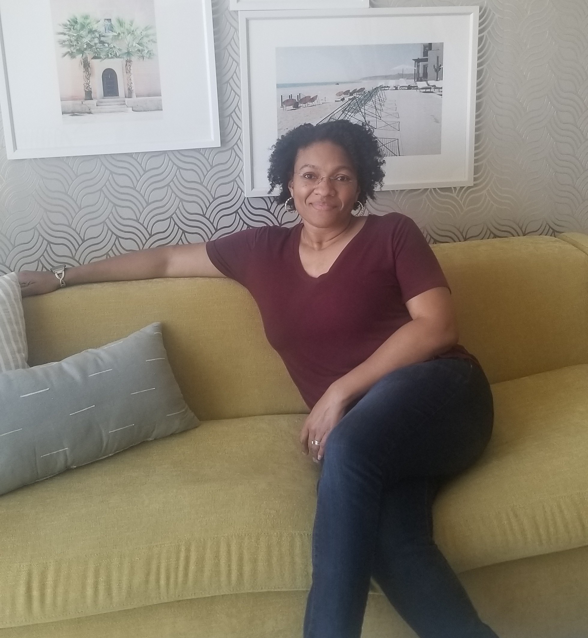 Woman sitting on a yellow sofa with a gray pillow, in a room with framed pictures on the wall behind her.