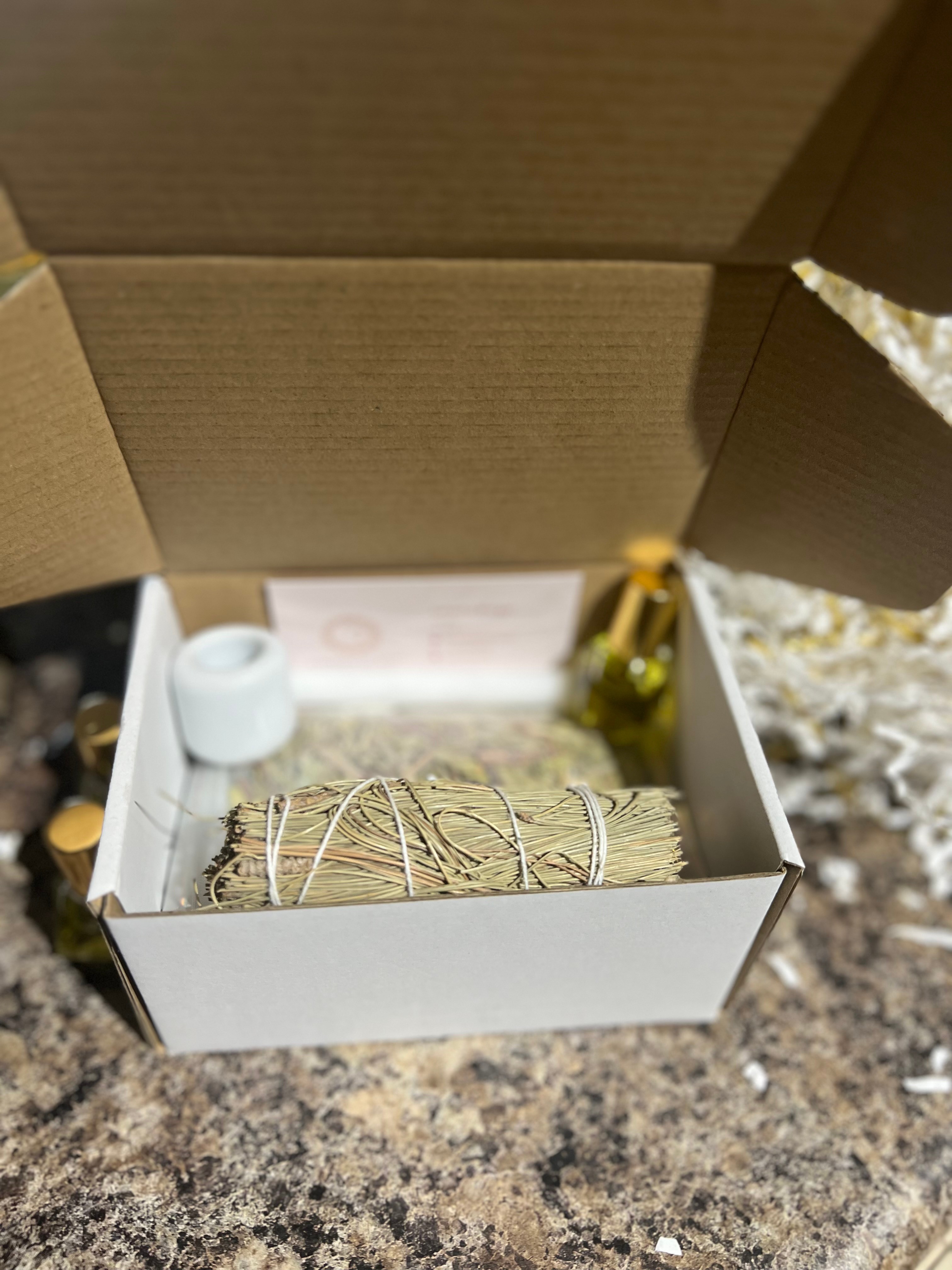 Open cardboard box containing a bundle of sage, a small white container, and a small yellow object, on a speckled surface.