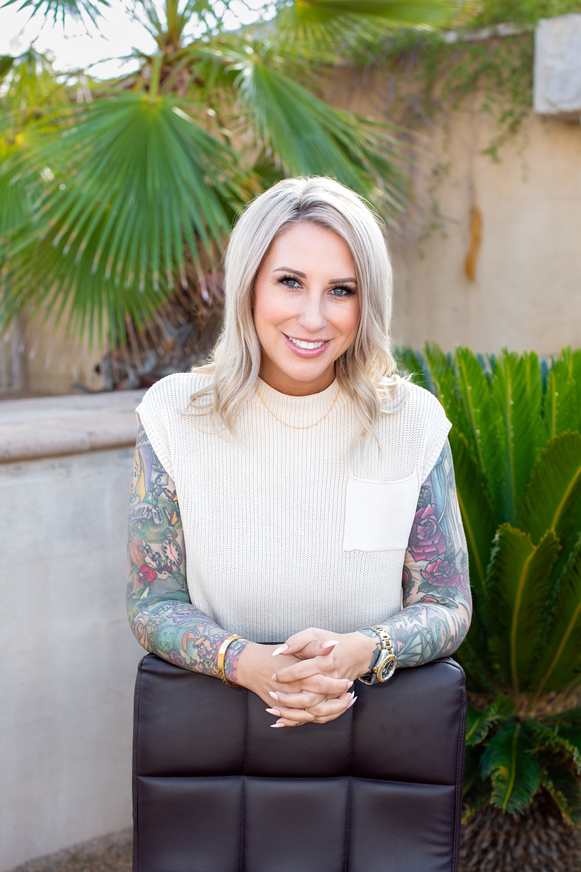 Smiling woman with blonde hair in a white sweater, standing outdoors with green plants and a wall behind her.