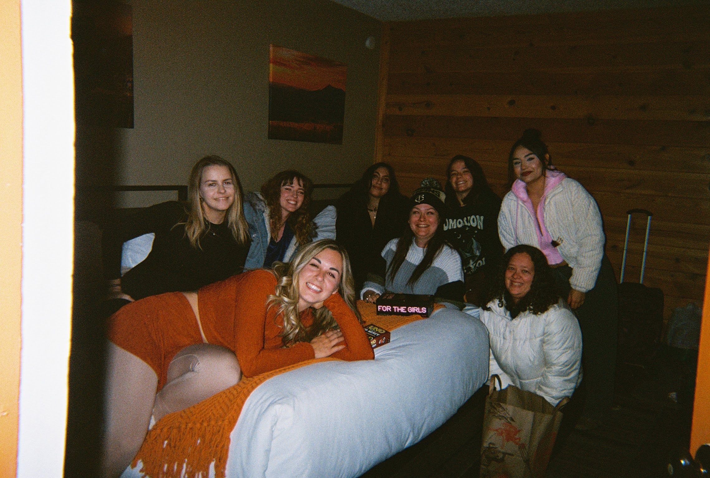 Group of nine women gathered in a room, some sitting on a bed and others standing, smiling at the camera.