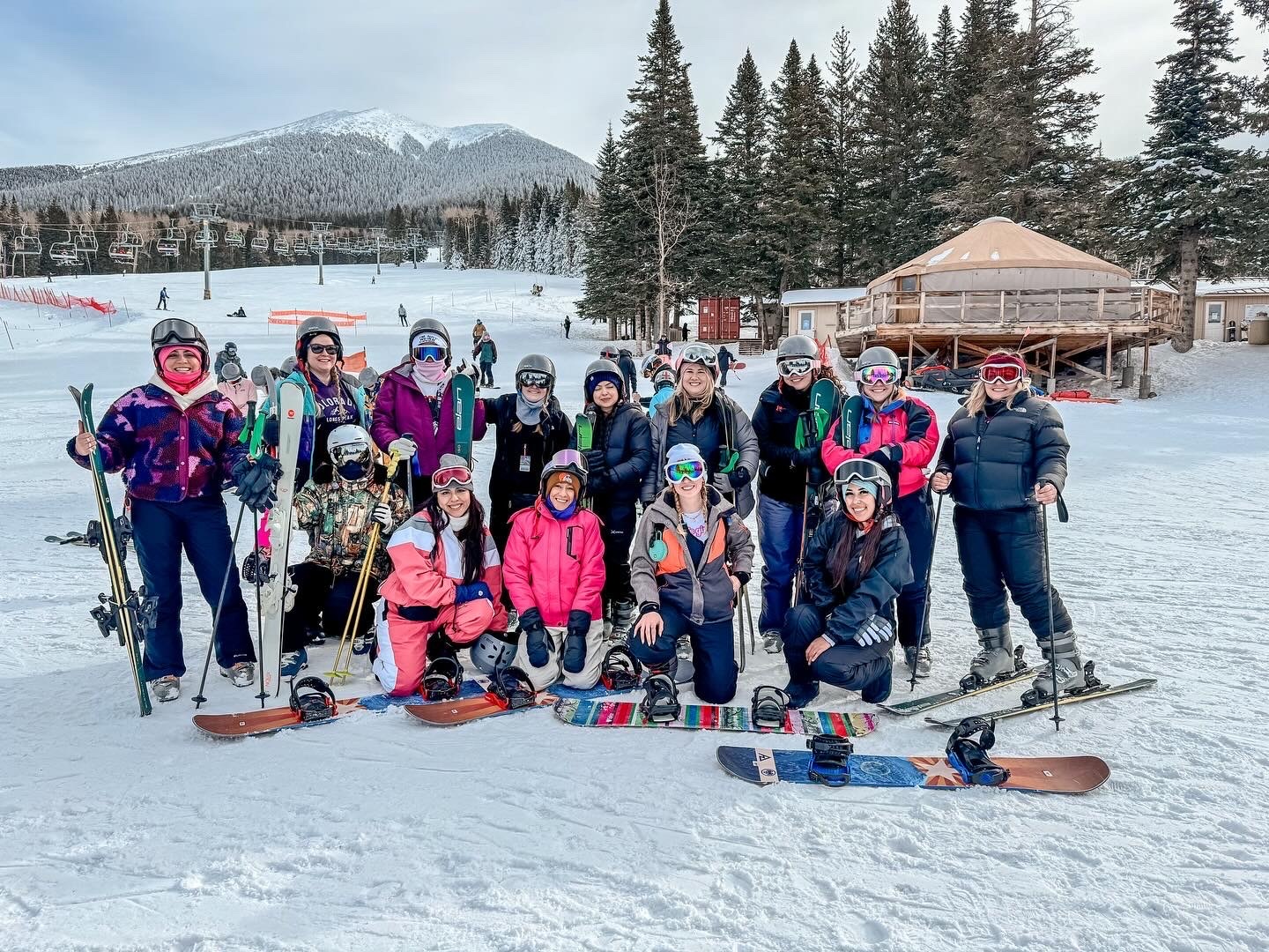 Group of people in winter clothing with skis and snowboards on snowy ground, trees, and a mountain in the background.