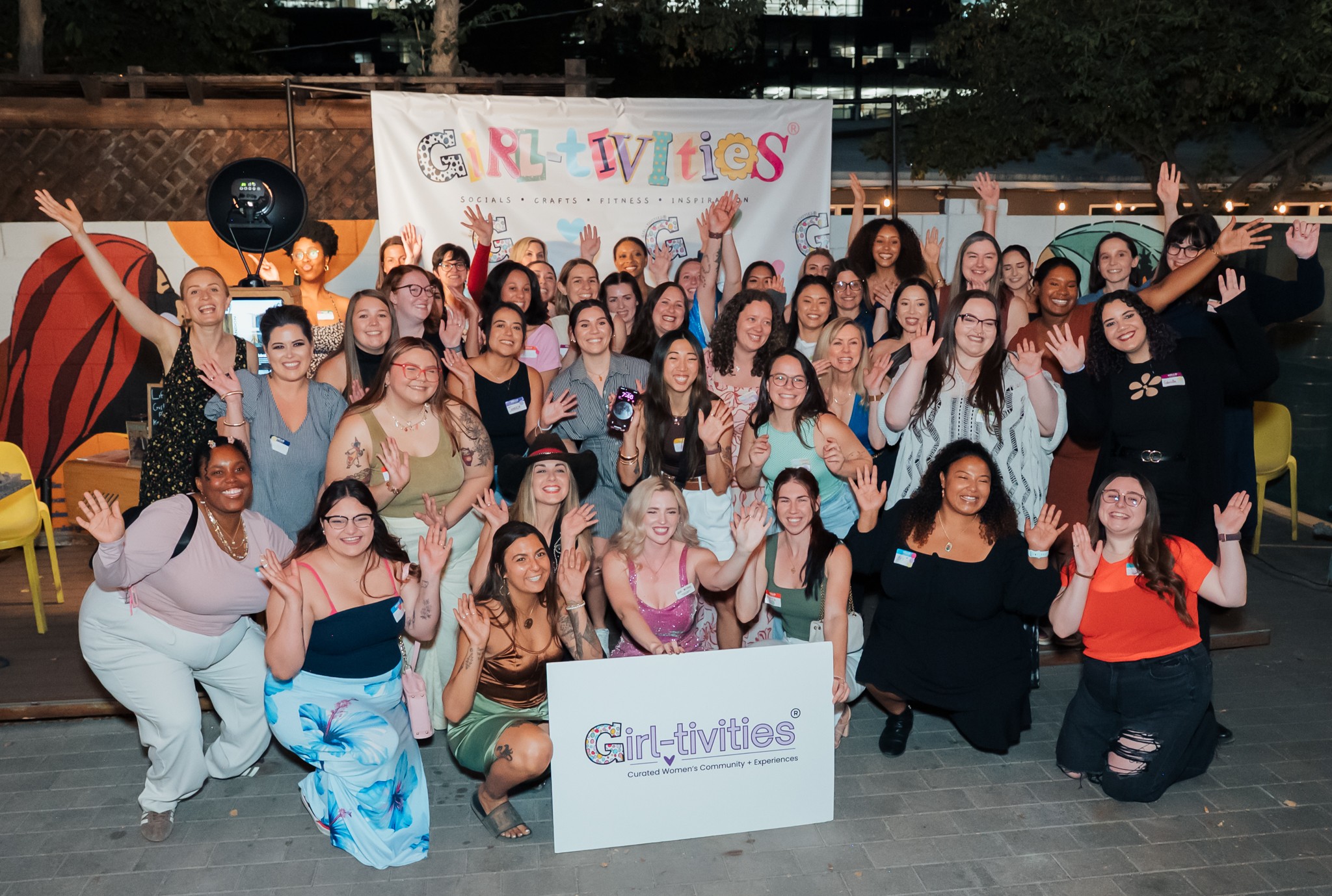 Group of women and girls smiling and raising hands outdoors, with banners and chairs in background.