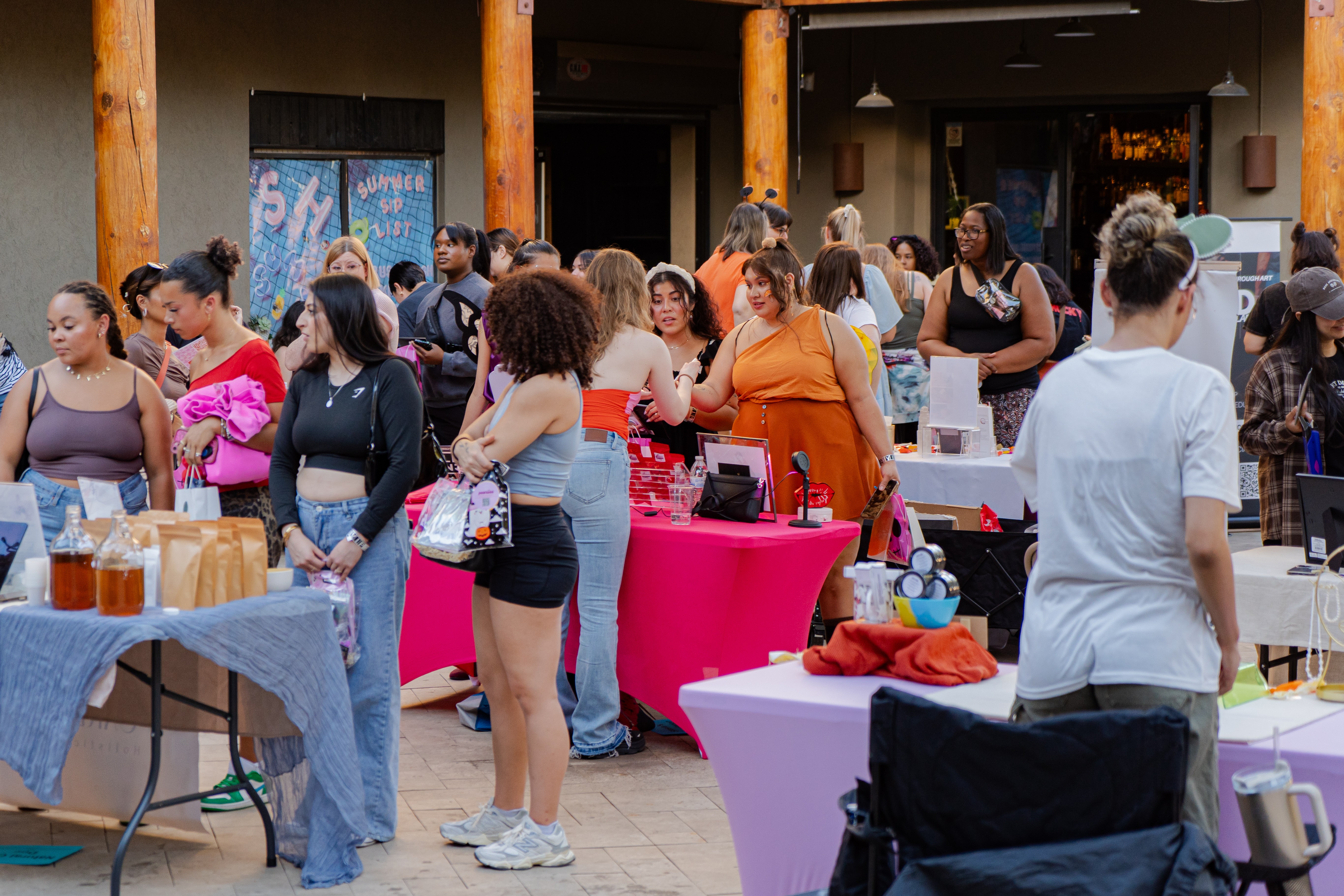 Group of people gathered around tables at an outdoor event, some browsing and others talking, with a building in the background.