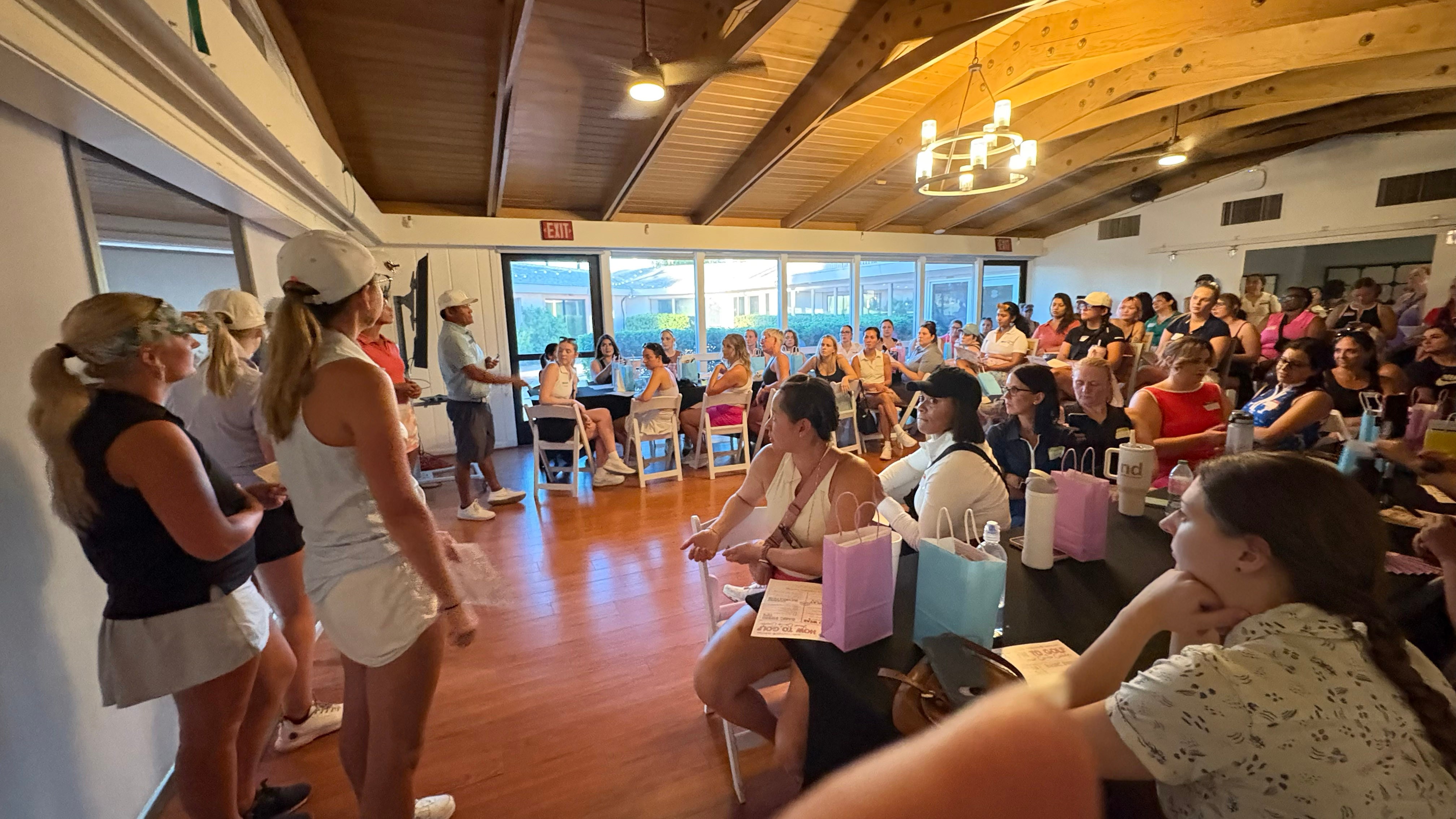 Group of women standing and sitting in a large room with wooden ceiling beams and large windows, some holding pink gift bags.