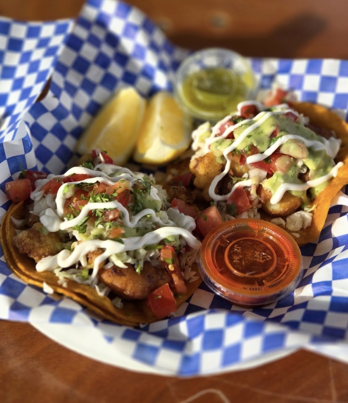 Two tacos with toppings, lemon wedges, and small containers of green and red sauces on checkered paper.