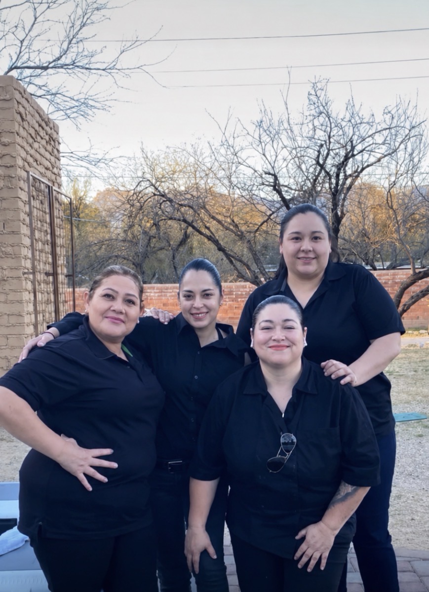 Four women standing outdoors, smiling, with trees and a brick wall in the background.