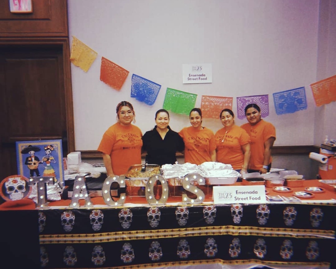 Five women stand behind a decorated table with food and drinks, colorful banners hang on the wall, skull-patterned tablecloth, and a sign reading 'Lemonade Street Food'.