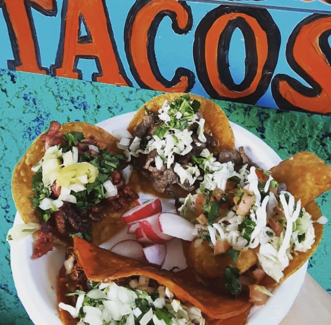Plate of tacos with meat, vegetables, and toppings, in front of a colorful sign that says 'TACOS'.