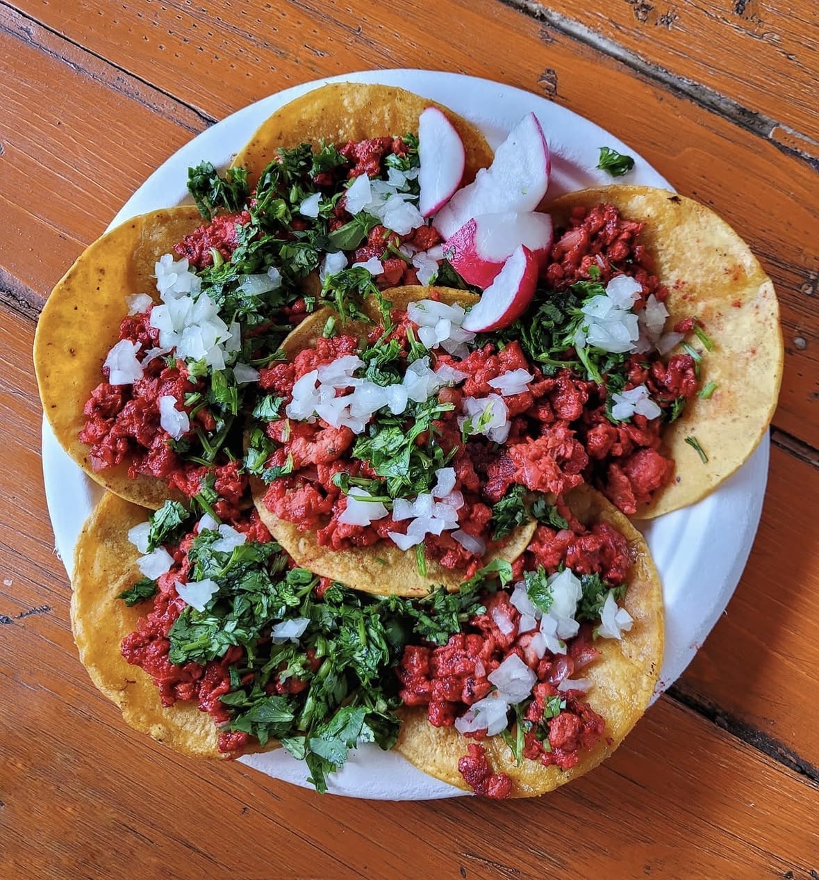 Plate with three tacos filled with meat, chopped onions, cilantro, and radish slices on a wooden table.