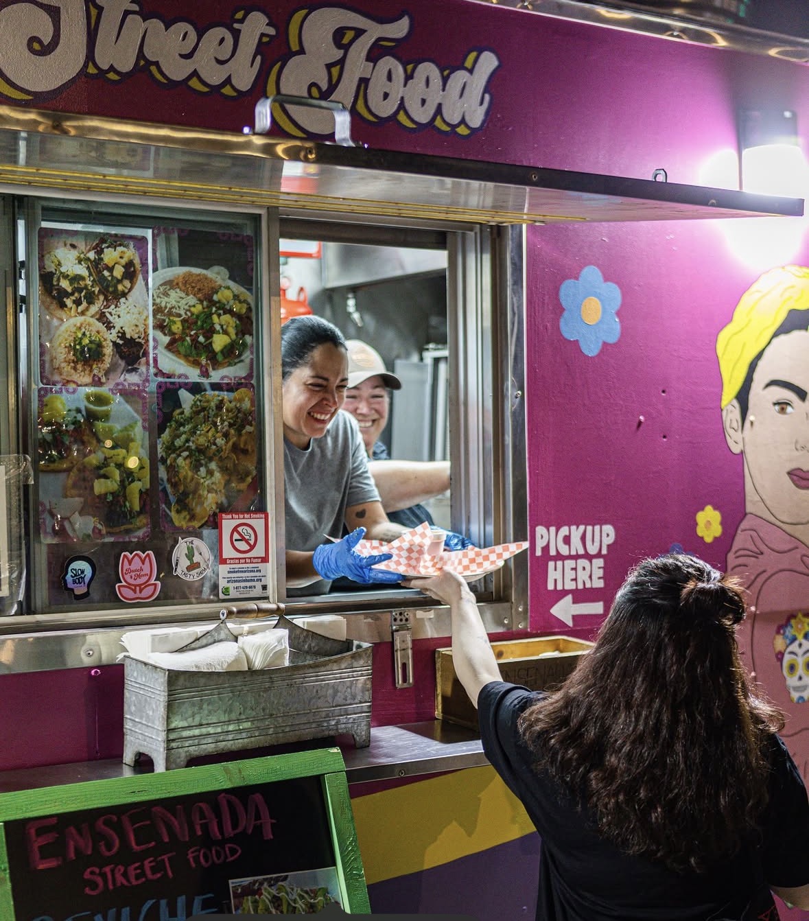 Woman with long dark hair reaching out to a smiling woman inside a colorful street food stall, with a menu display and bright pink wall.