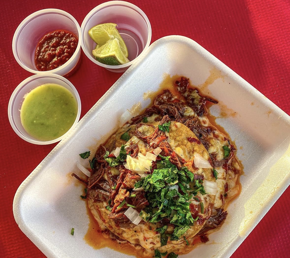 Plate with a dish topped with herbs, surrounded by three small bowls of sauces and lime wedges, on a red surface.
