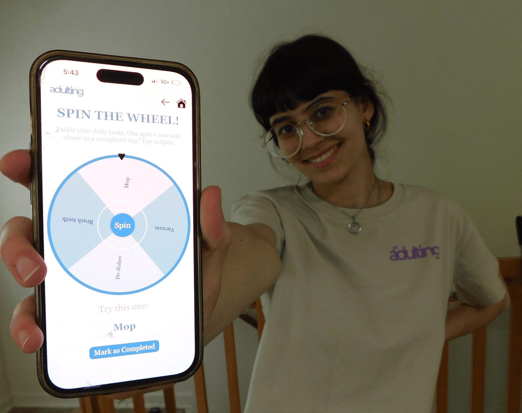 Young woman with glasses smiling, holding a smartphone showing a spinning wheel app, standing indoors against a plain wall.