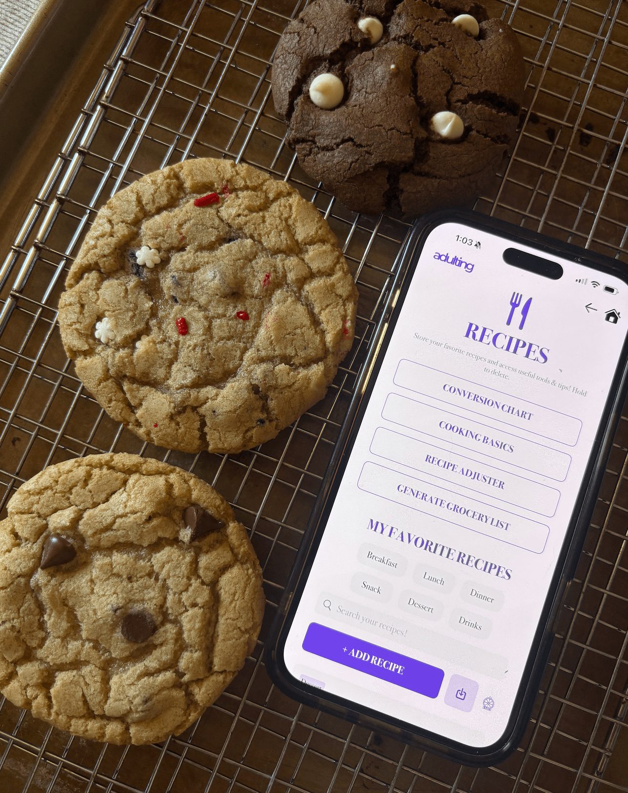 Three cookies on a cooling rack with a smartphone displaying a recipes app, all viewed from above.