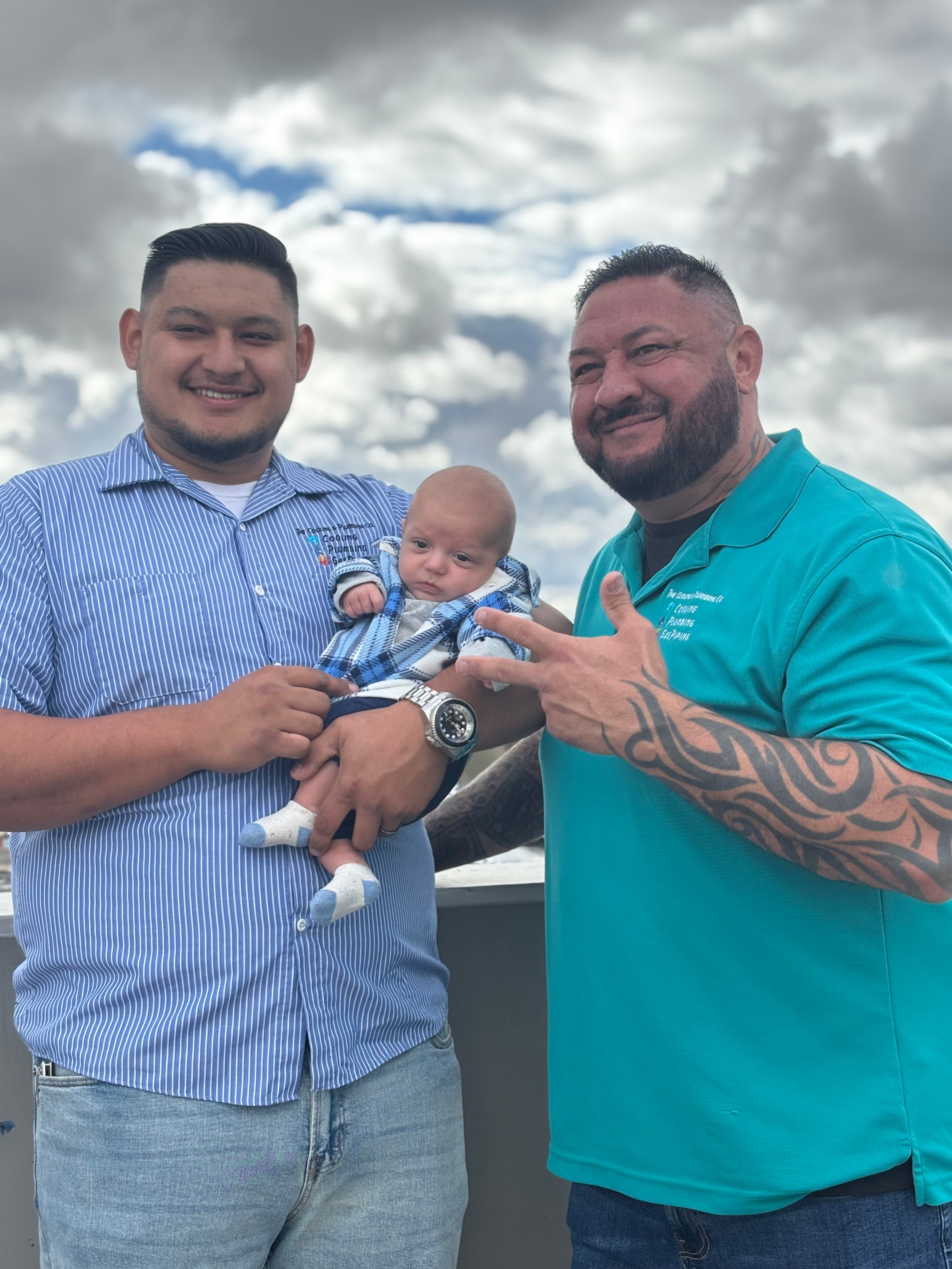 Two men holding a baby outdoors with cloudy sky background, smiling, one pointing at the baby, casual clothing, one with tattoos.