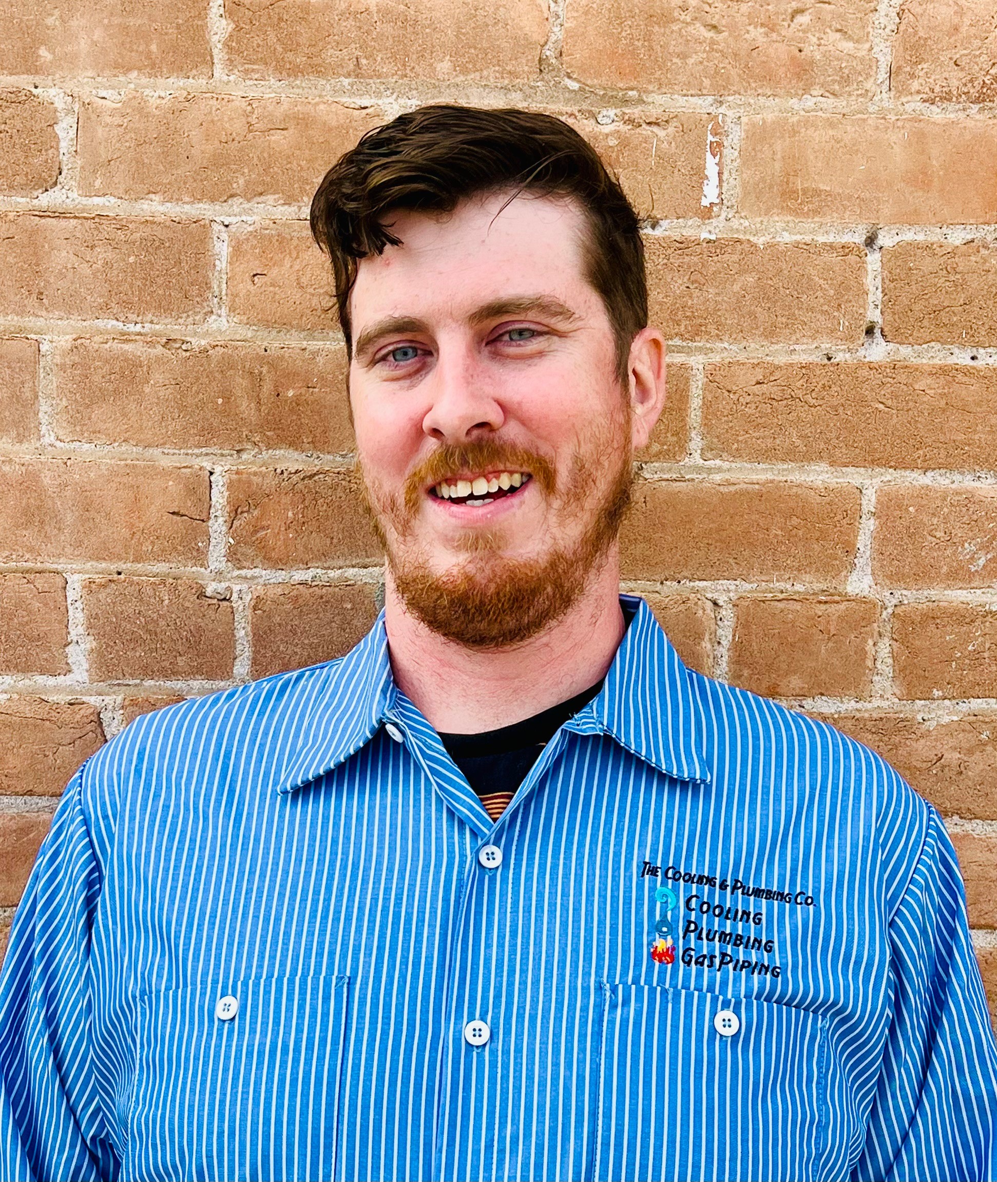 Young man with dark hair and beard smiling, wearing a blue striped shirt, standing against a brick wall.