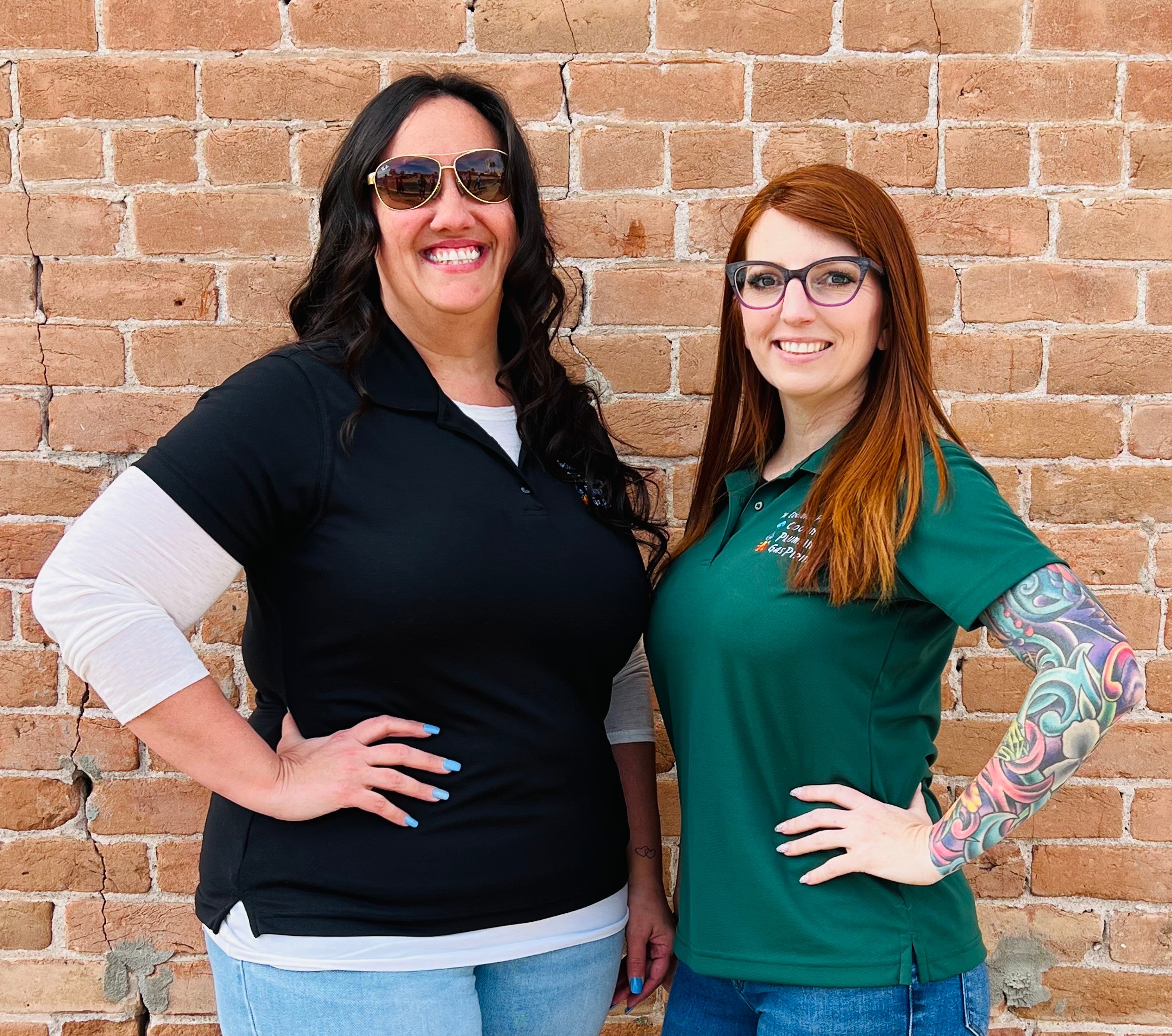 Two women stand side by side in front of a brick wall, smiling, with one wearing sunglasses and the other glasses.