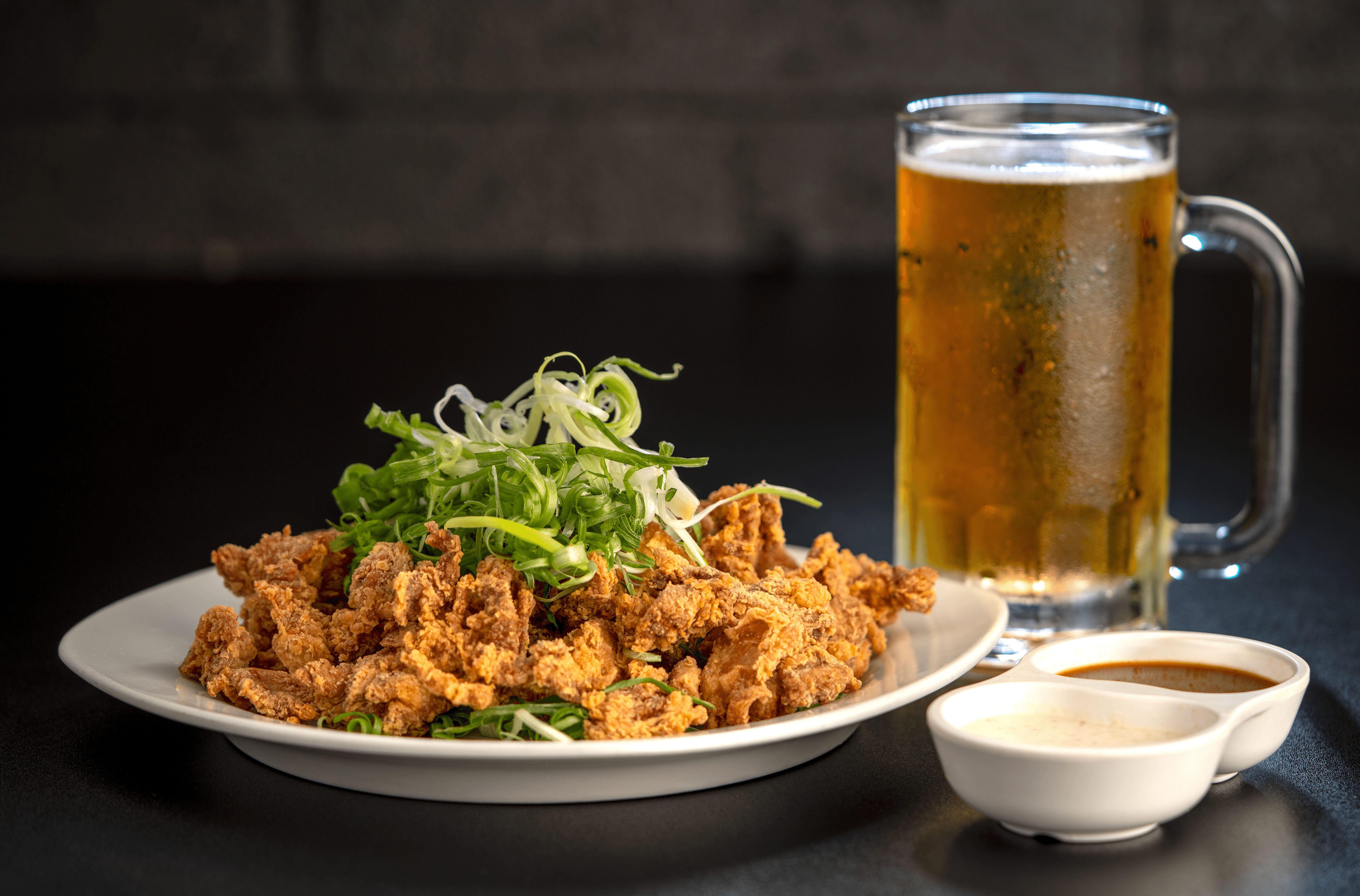 Plate of fried food with green garnish, a glass of beer, and two small bowls of sauce on a dark surface.