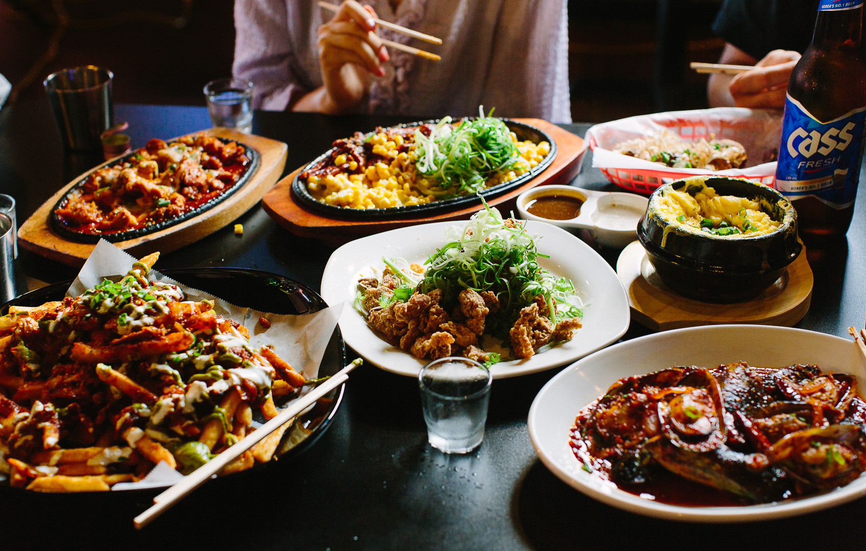 Various dishes of Asian cuisine on a table, including fried rice, noodles, and grilled meat, with people eating in the background.