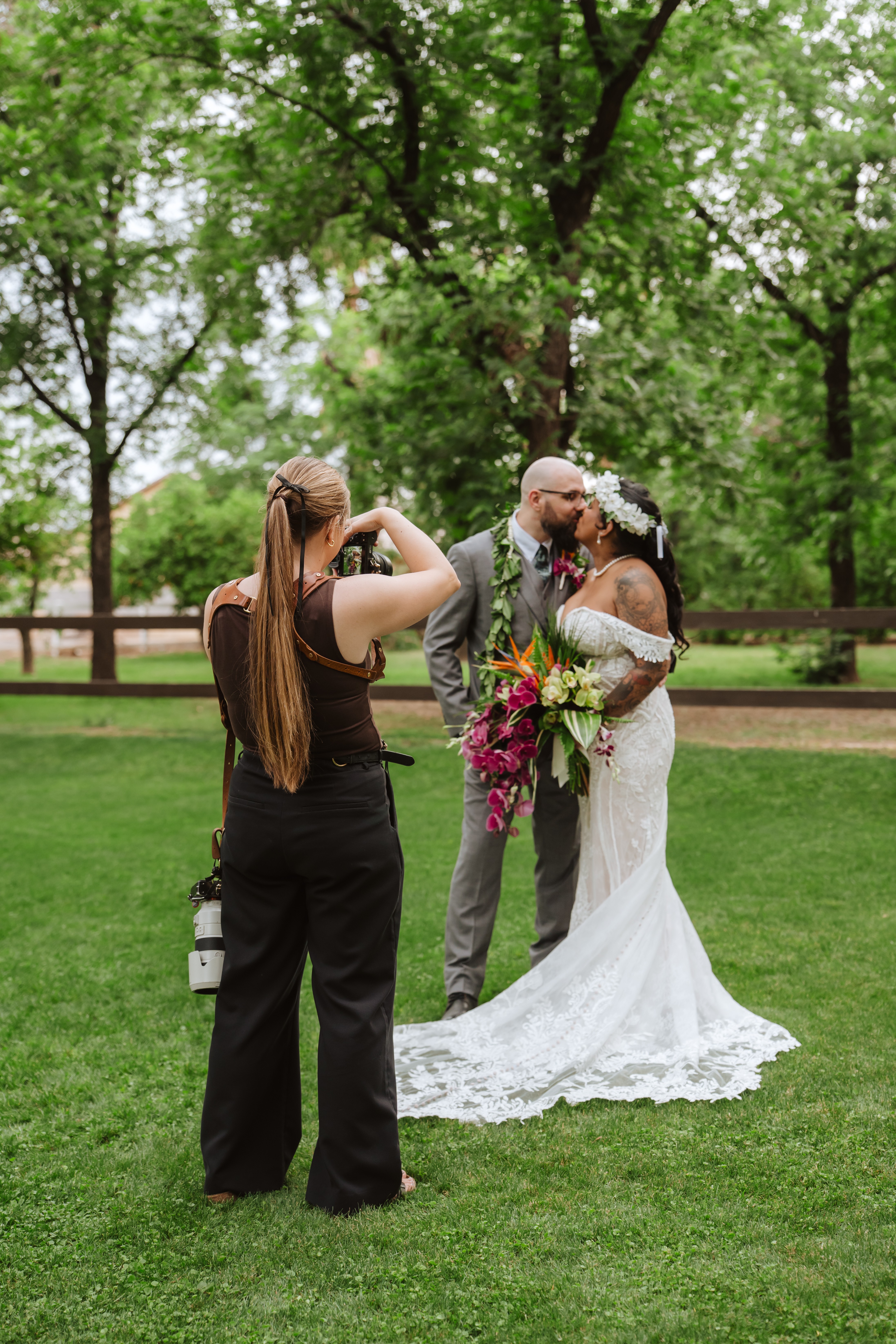 Bride and groom kiss outdoors, photographer captures moment, bride holds bouquet, trees and fence in background, grassy area.