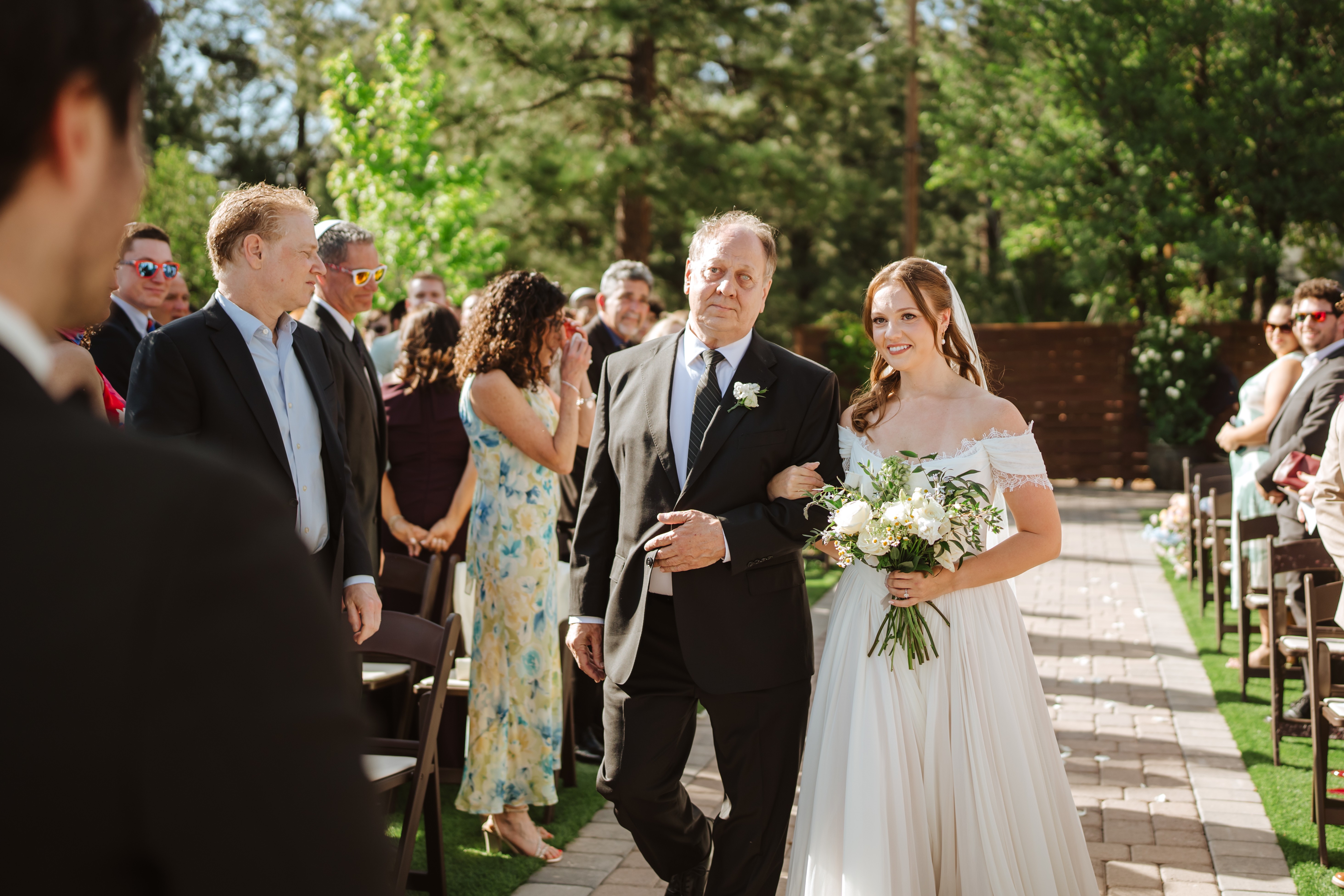 A bride in a white dress holding a bouquet walks down an outdoor aisle with a man in a suit, surrounded by guests.
