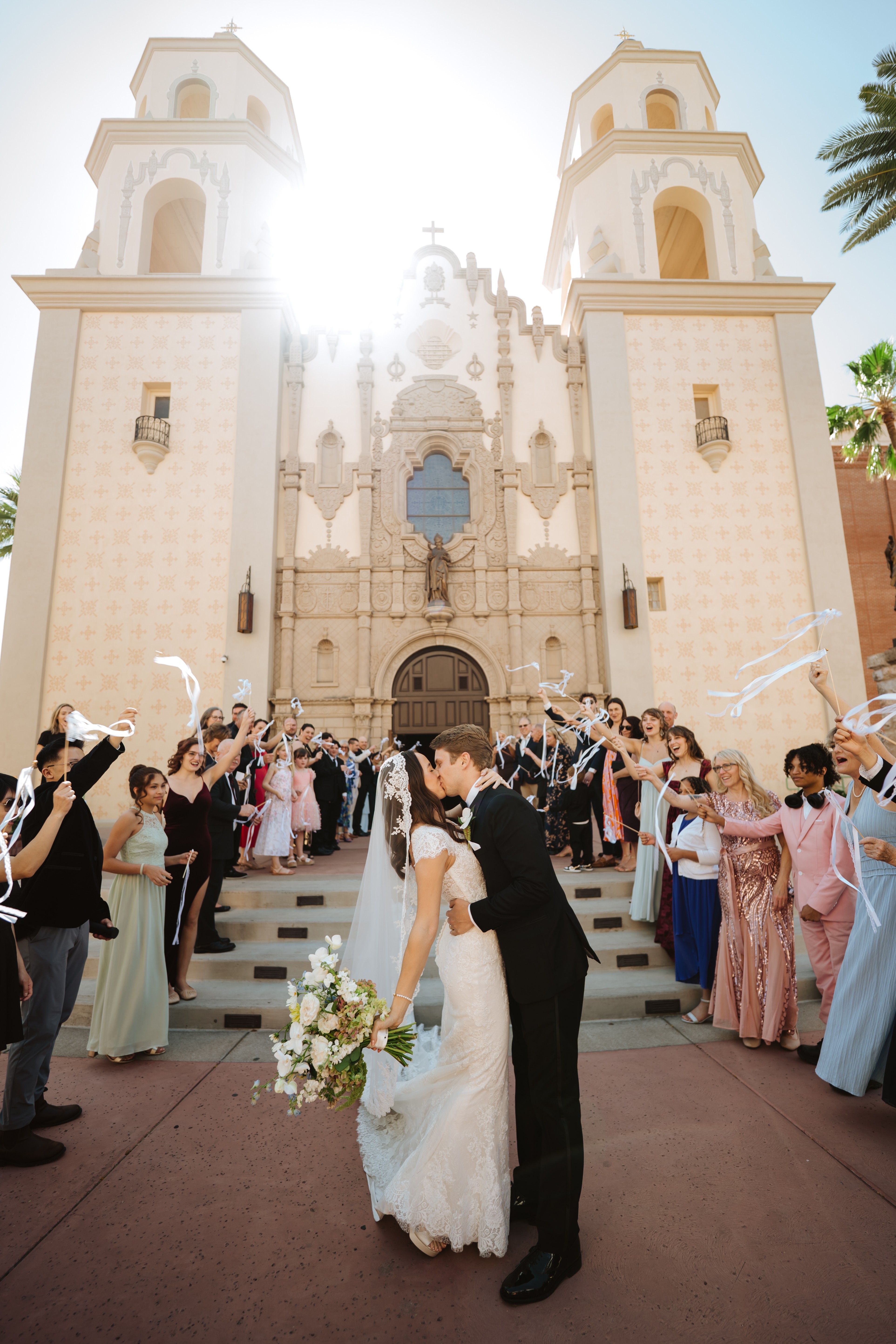 Bride and groom kiss in front of a church with wedding guests celebrating around them, sunlight shining behind the church.
