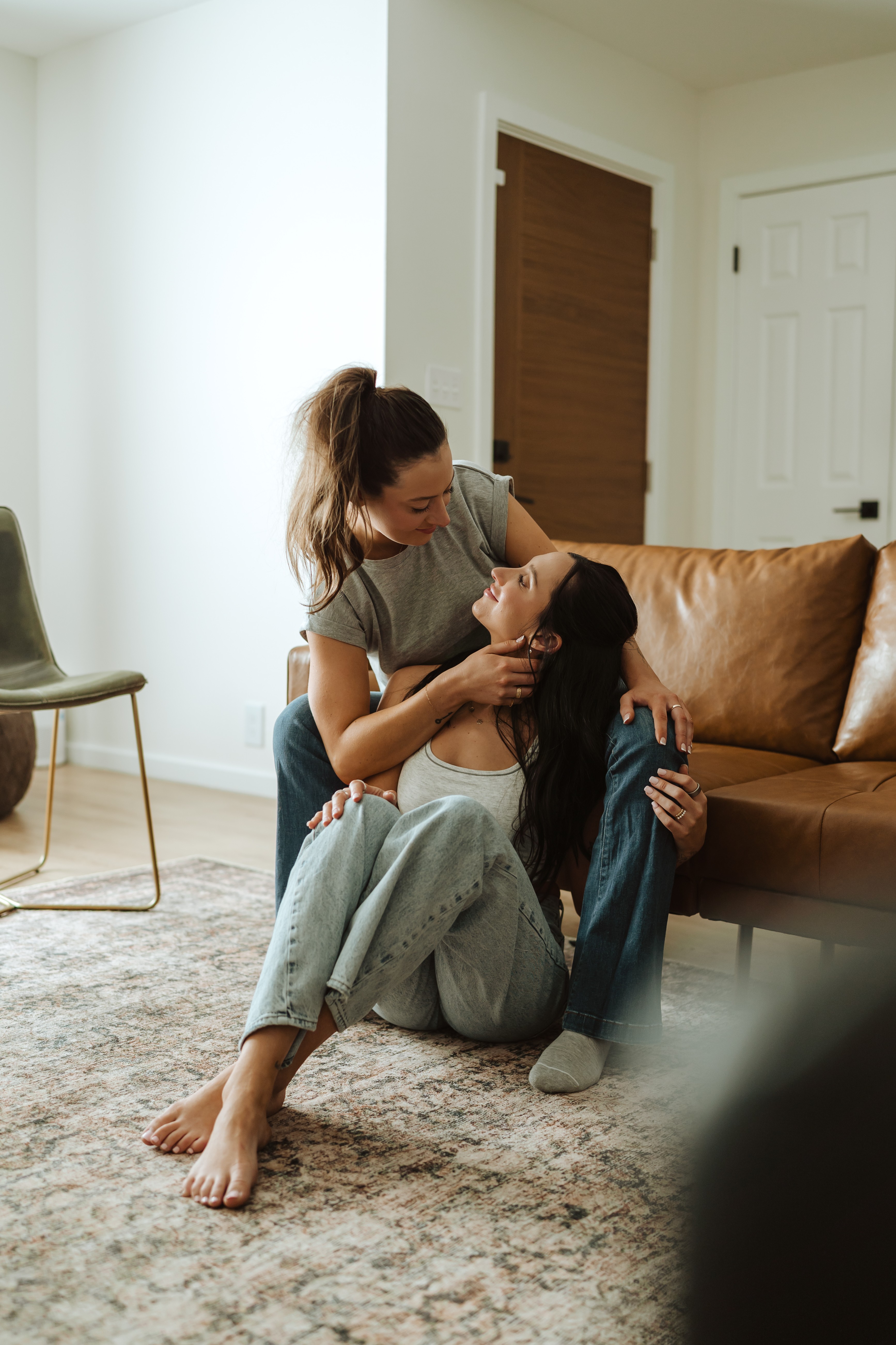 Two women sit on the floor, one holding the other's face, in a living room with a brown sofa and a white wall.