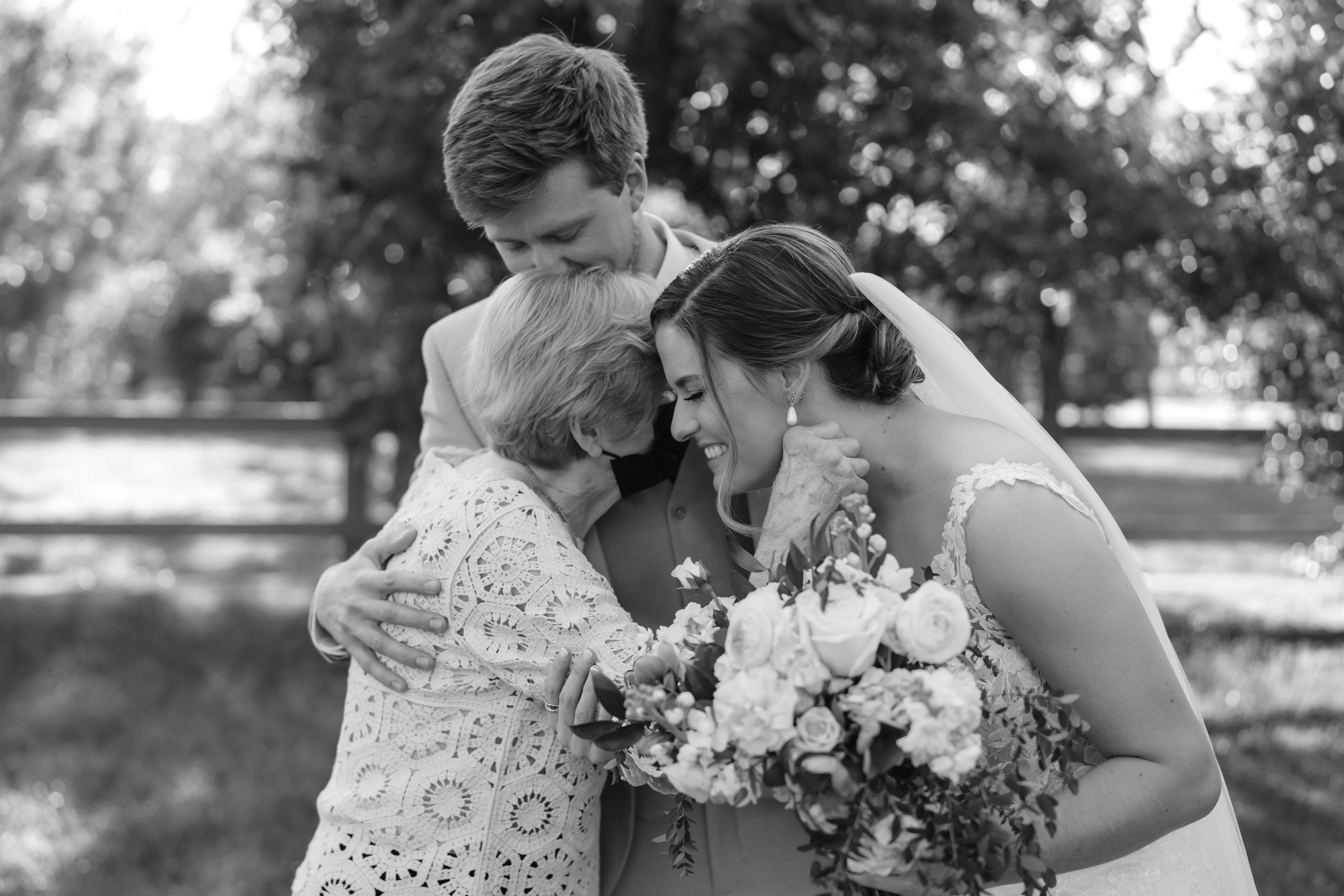 Four people, including a bride holding a bouquet, embrace outdoors with trees in the background.
