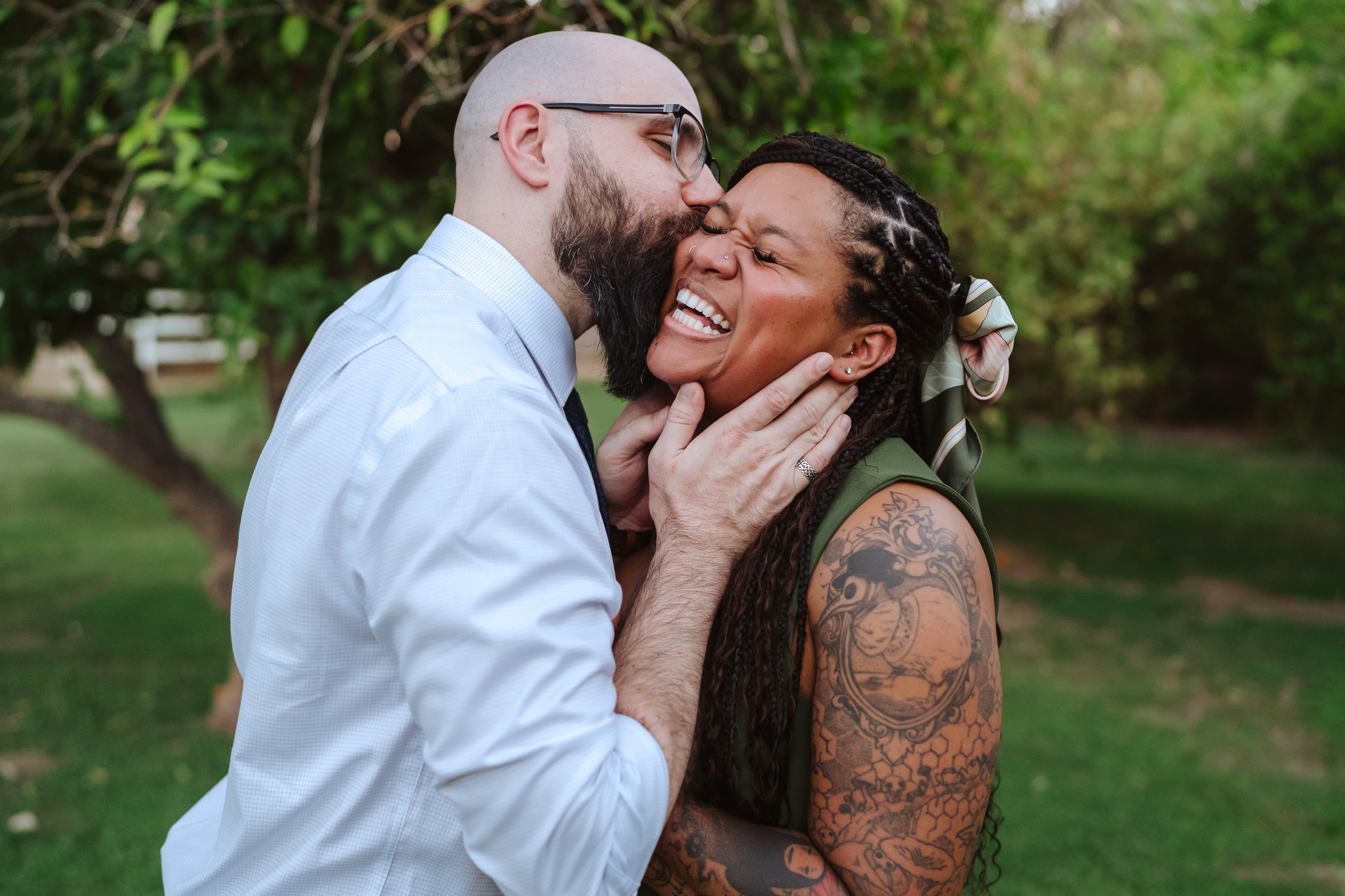 Two people smiling and laughing outdoors, one with glasses and a beard, the other with tattoos and long hair, embracing.