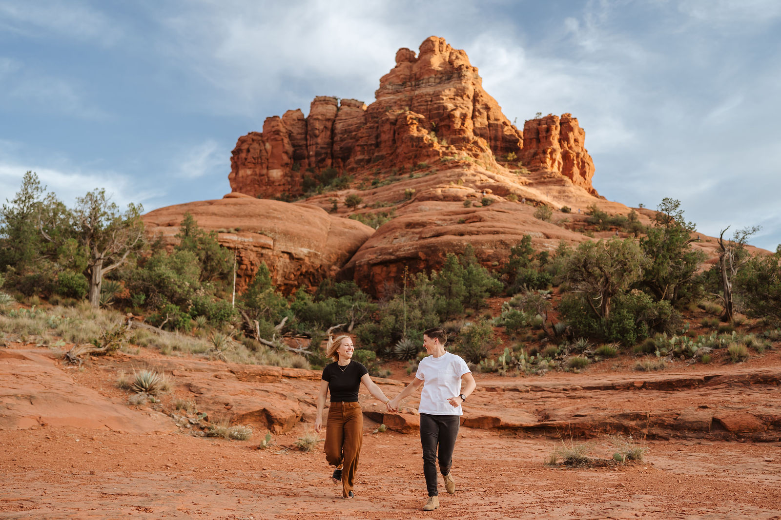 Two people walk hand in hand in a desert landscape with red rock formations and sparse vegetation under a partly cloudy sky.