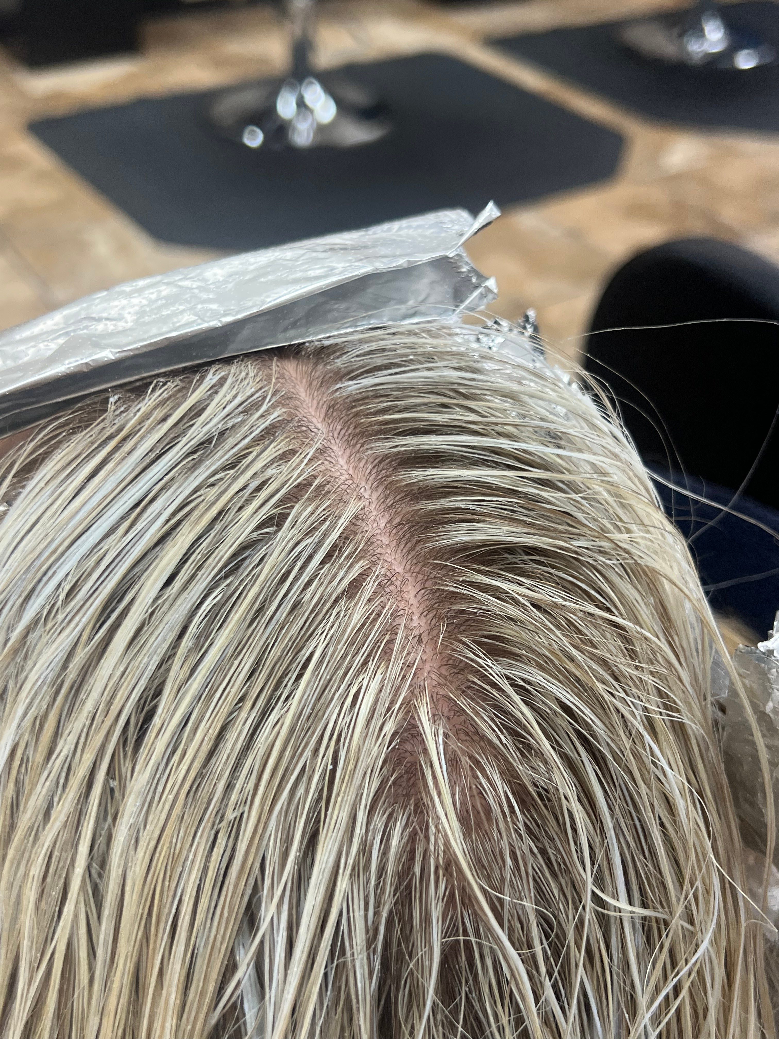 Close-up of a person's scalp and hair, showing blonde hair with roots visible, hair parted to the side.