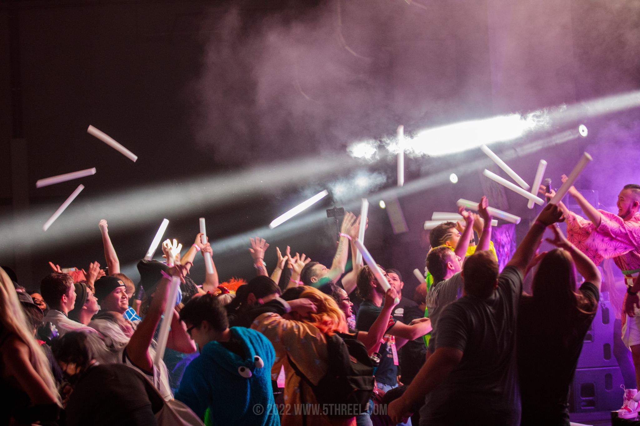 Crowd at a concert with hands raised, colorful lighting, and stage smoke, enjoying the performance.