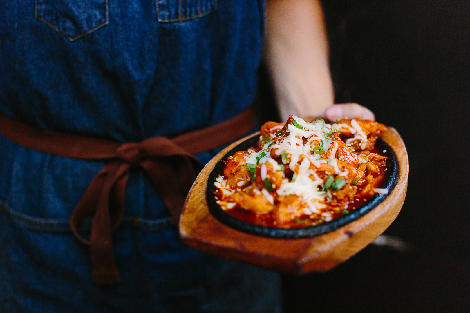 Person holding a wooden bowl of pasta with cheese and herbs, wearing a blue apron and brown belt, dark background.