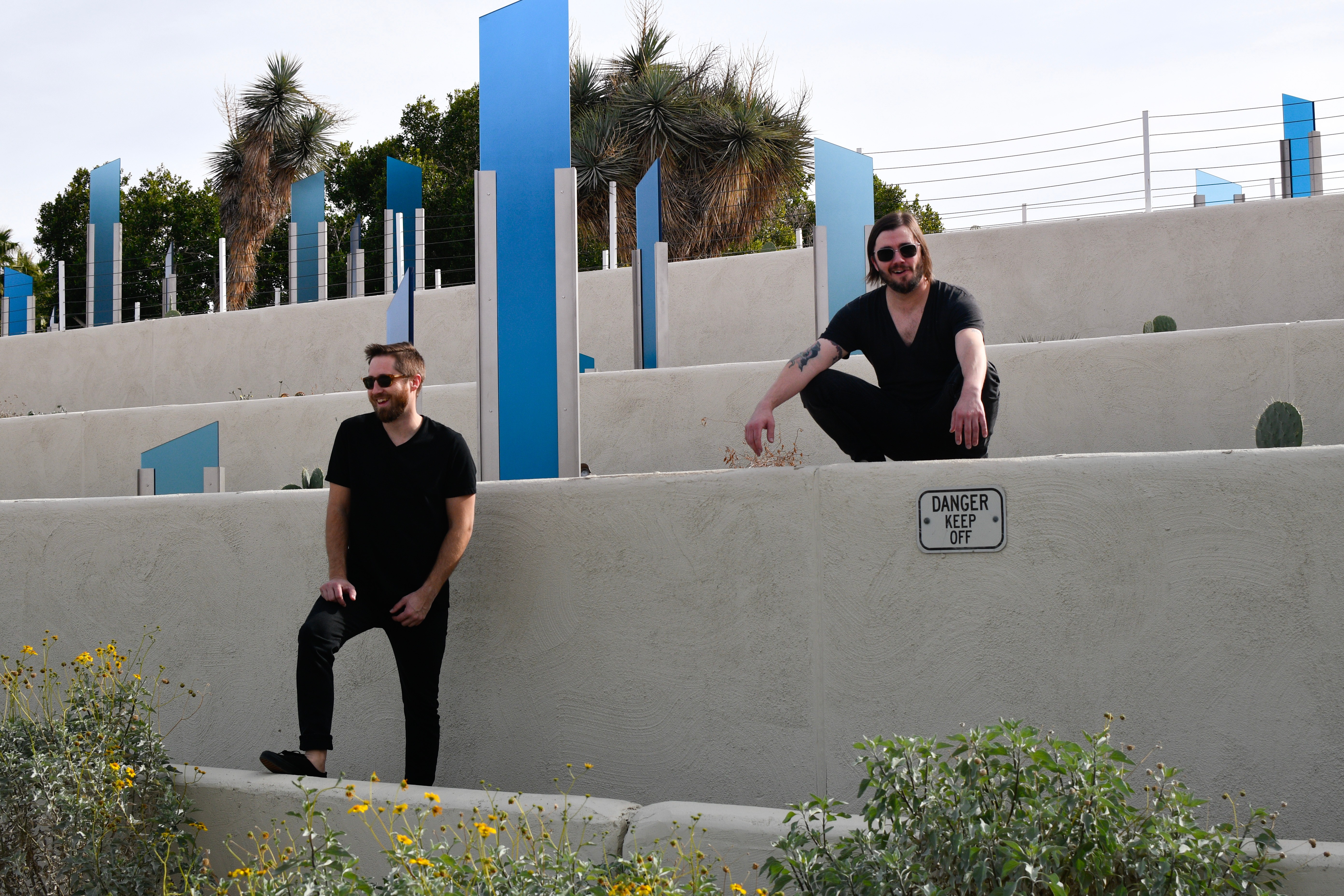 Two men sit on concrete steps outdoors, with blue vertical structures and palm trees in the background, under a partly cloudy sky.