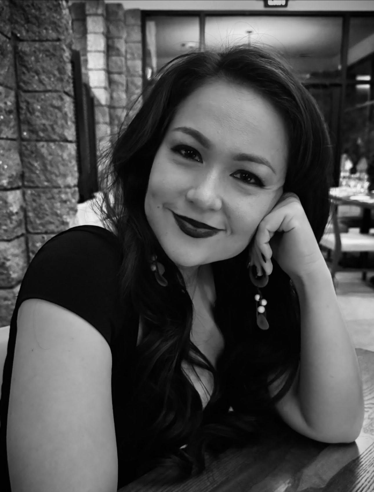 Woman with long dark hair and dark lipstick resting her chin on her hand, smiling at the camera, in a cafe setting.
