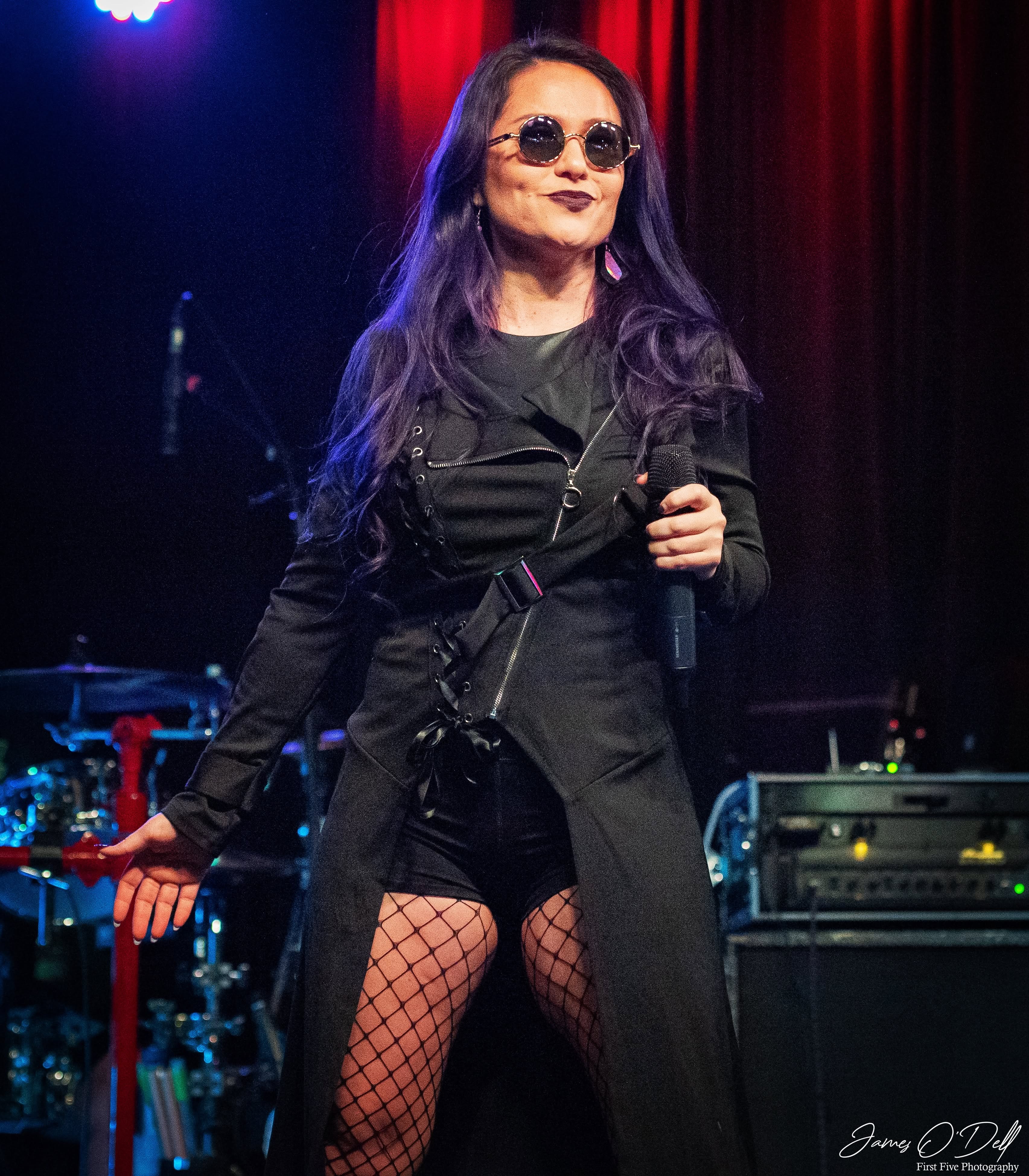 Woman with long purple hair wearing sunglasses, black outfit, fishnet stockings, holding a microphone on stage.