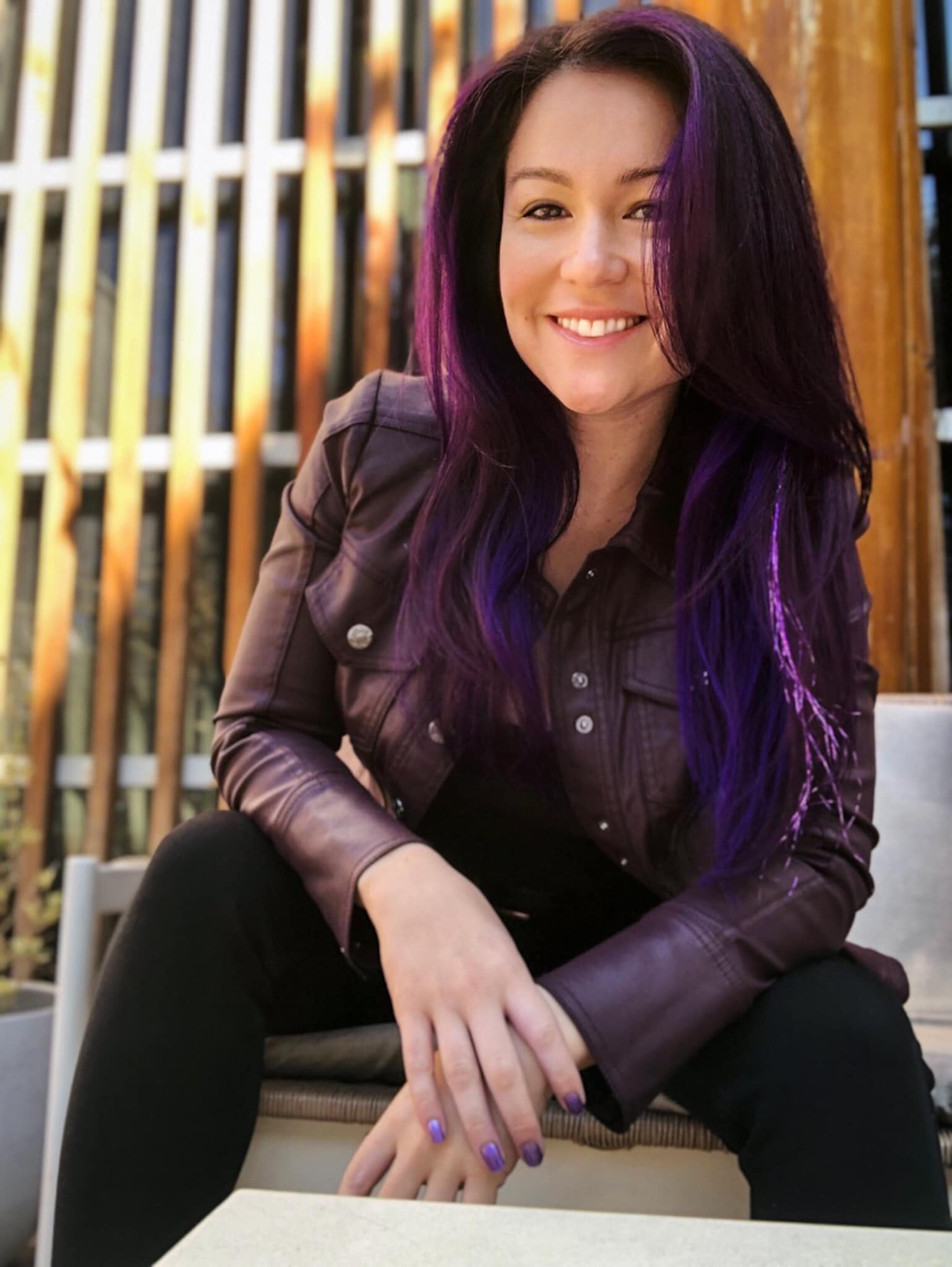 Smiling woman with long purple hair, sitting outdoors near a wooden fence, wearing a dark jacket and black pants.