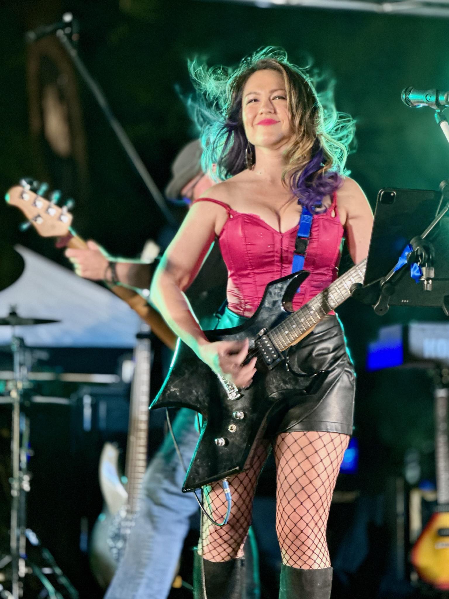 Woman playing electric guitar on stage, smiling, with green lighting, wearing a pink top, black shorts, and fishnet stockings.