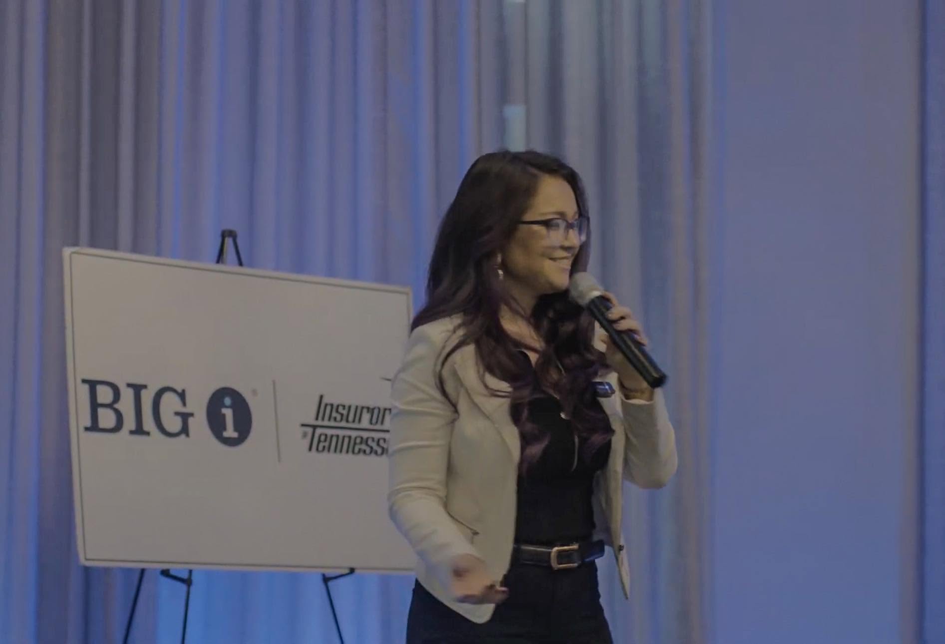 Woman speaking into a microphone next to a presentation screen with text, in a room with blue curtains.