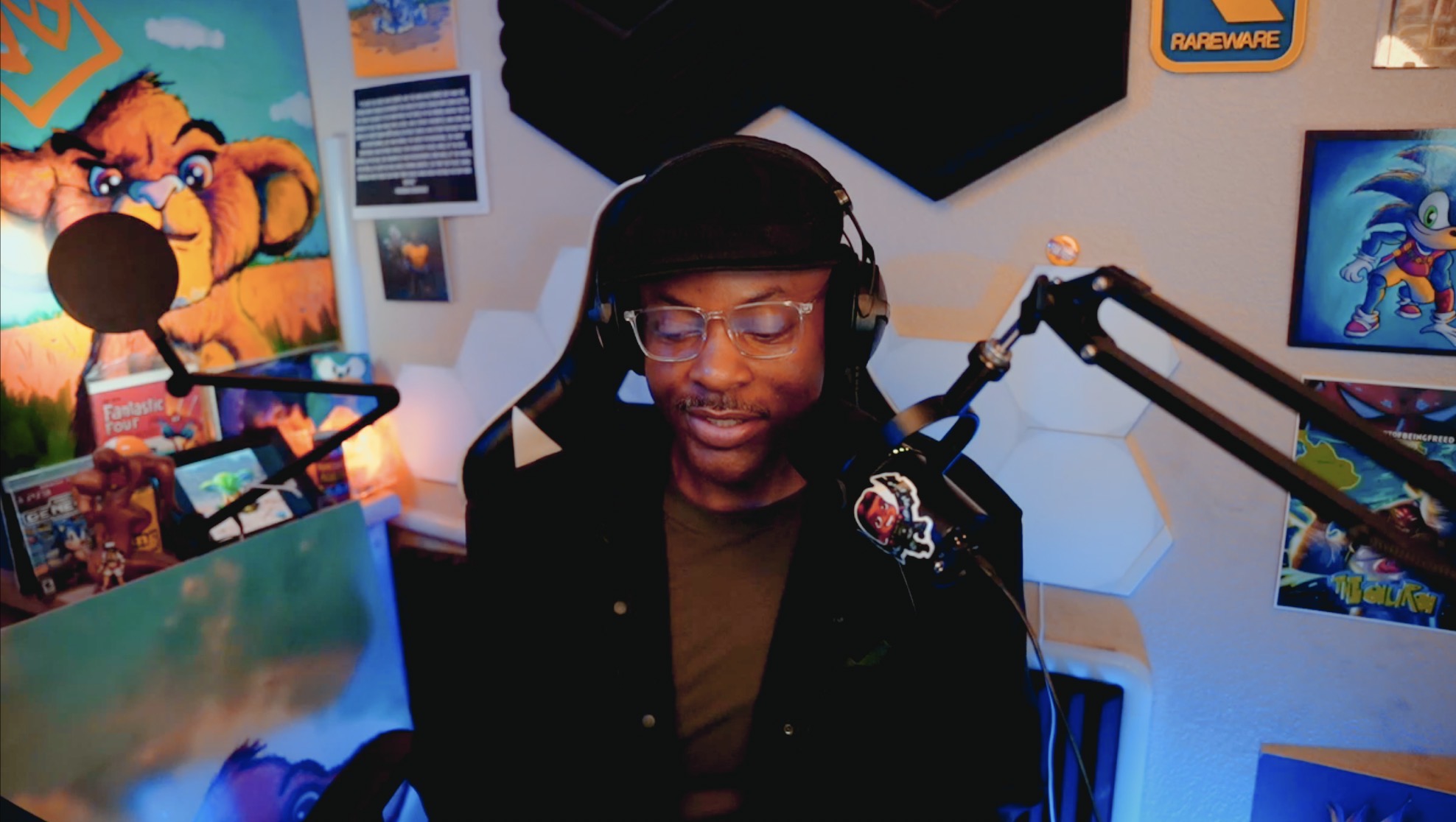 Person with glasses and headphones speaking into a microphone in a colorful room with posters and screens