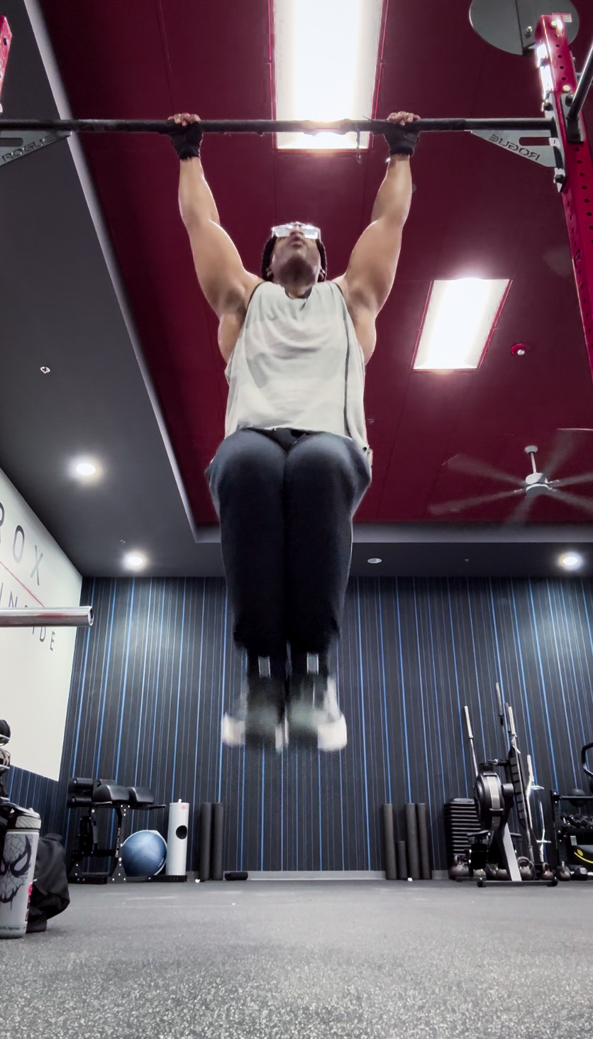 Person lifting a barbell overhead in a gym, hanging from a pull-up bar, with gym equipment in the background.