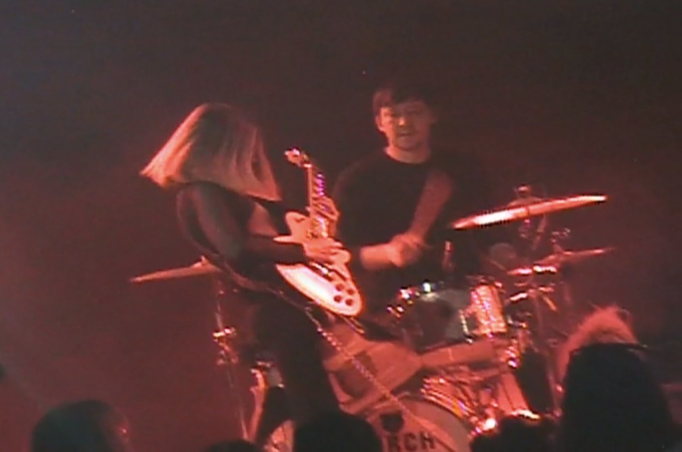 Two musicians perform on stage with guitars and drums, with a dark background and red lighting.