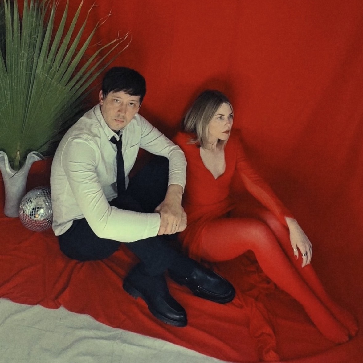 A man and woman sit on a red fabric surface with a red background, surrounded by green leaves and decorative objects.