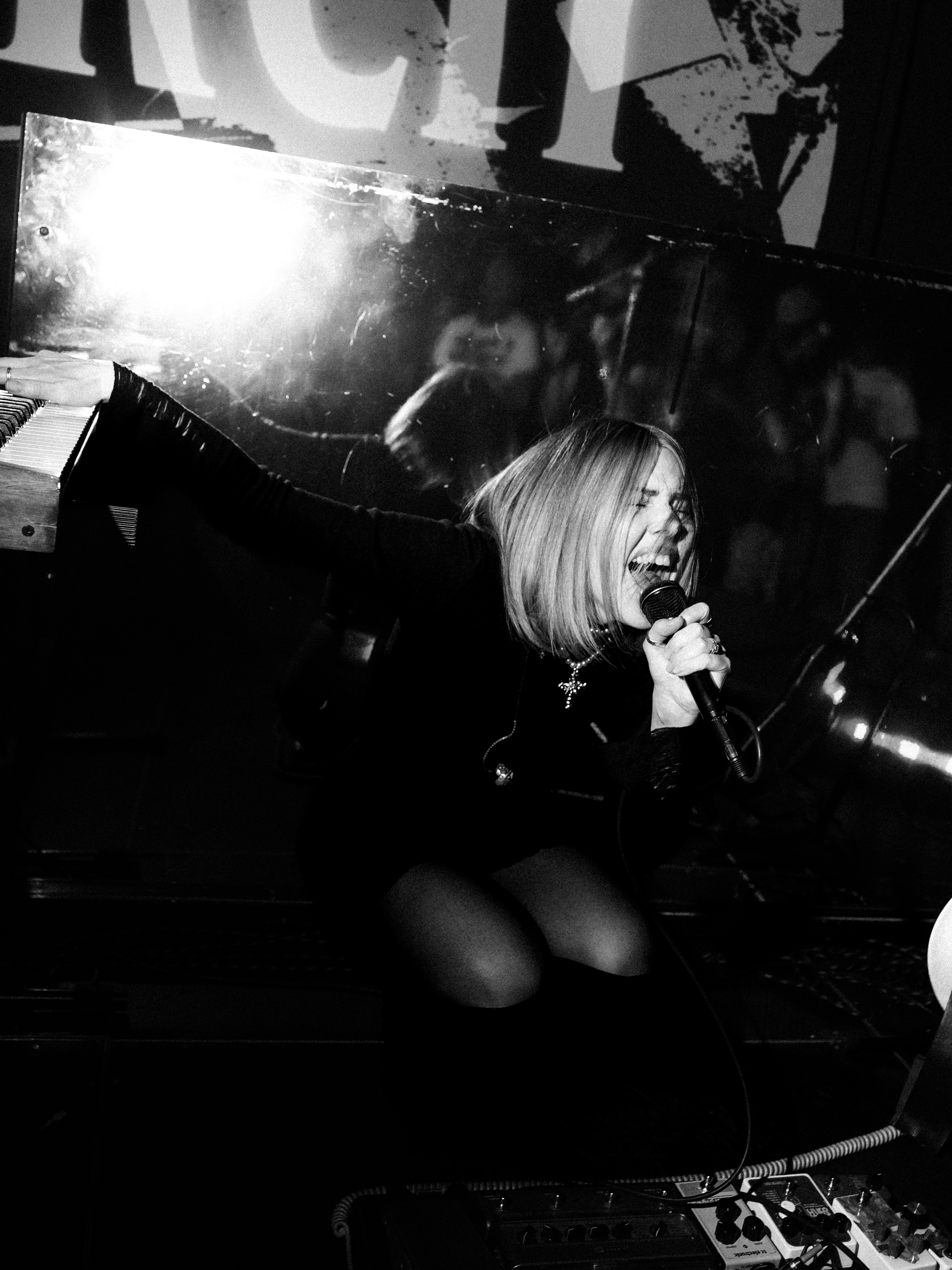 A woman with short blonde hair singing into a microphone on stage, with a reflective background and audience in the distance.