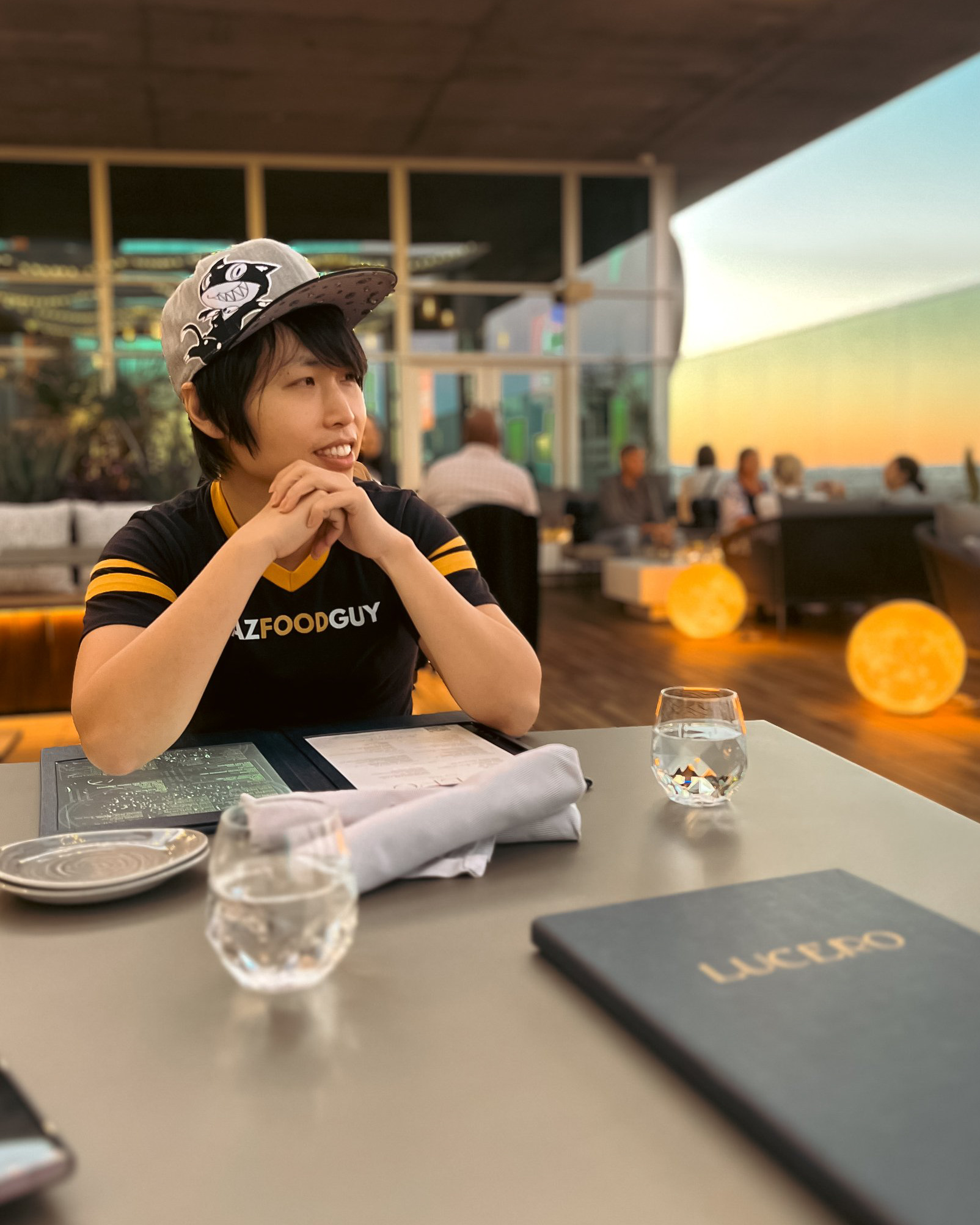 Young woman wearing a hat and black shirt sitting at a table with water glasses, smiling, with a sunset view outside.