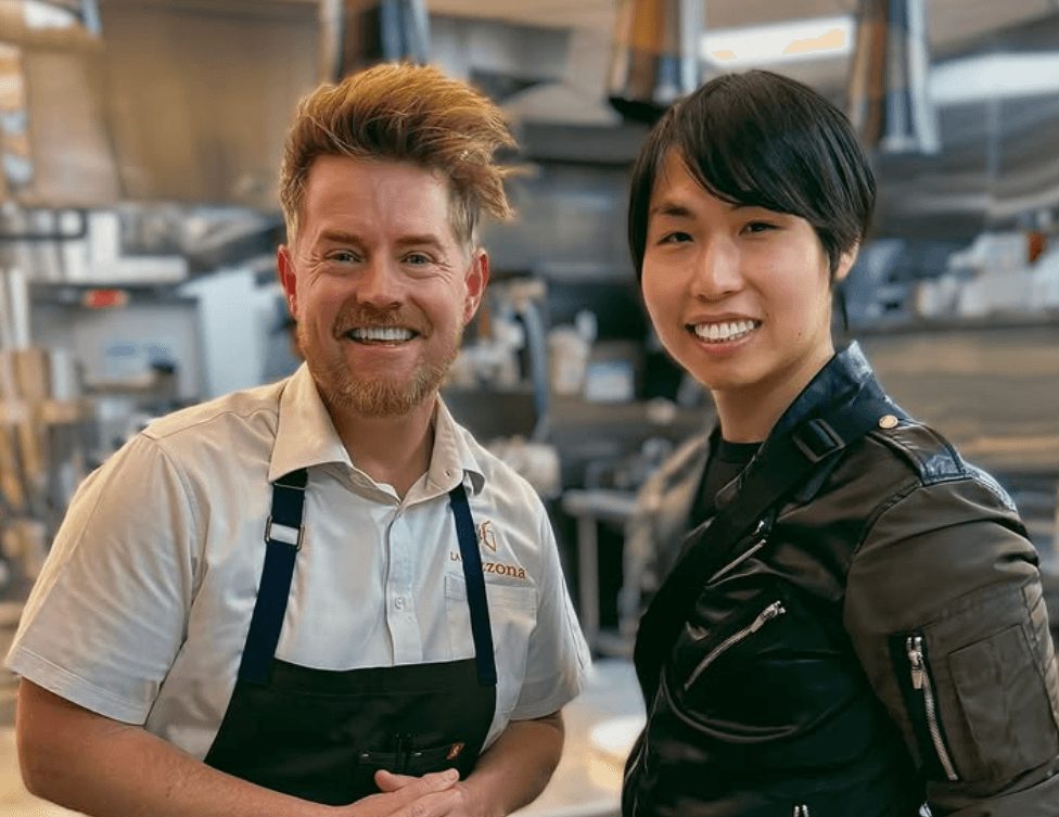 Two people smiling in a kitchen, one with a beard and apron, the other with short black hair and a black jacket.