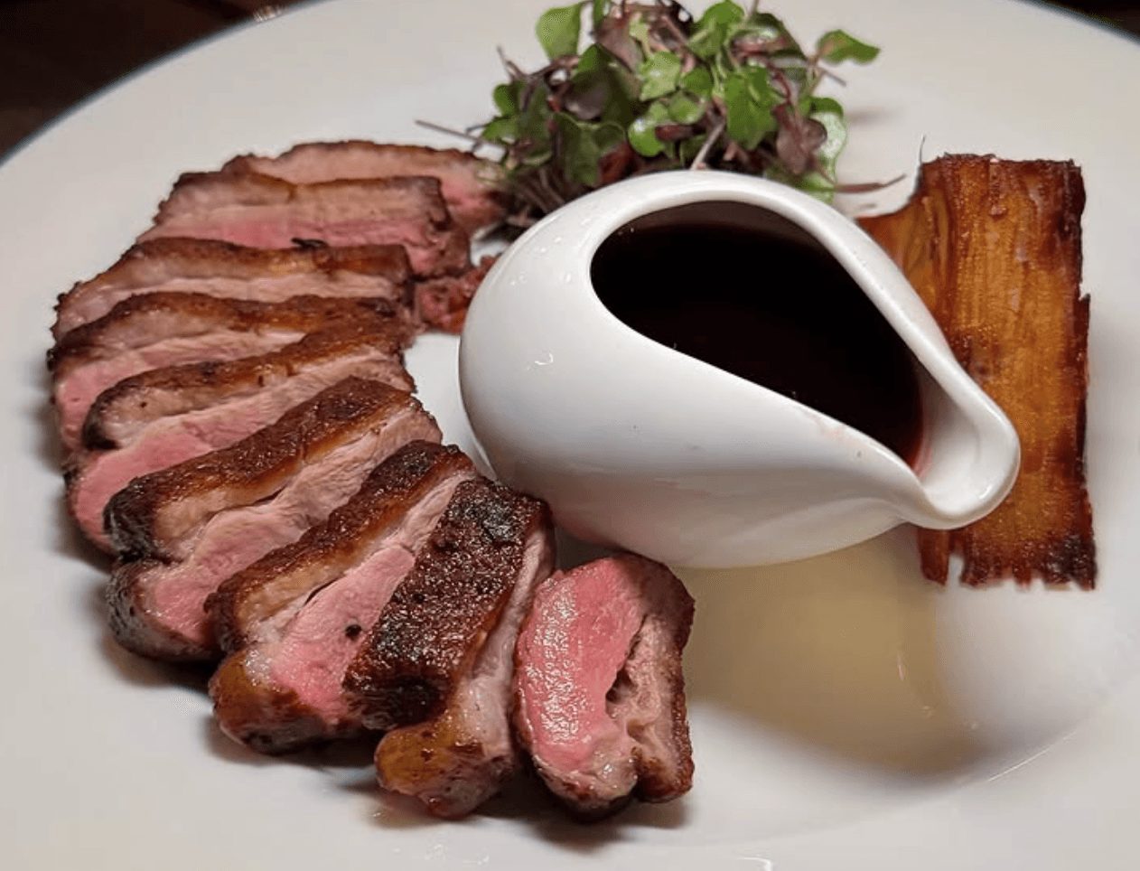 Sliced steak with sauce in a white pitcher, side of greens, and a piece of roasted potato on a white plate.