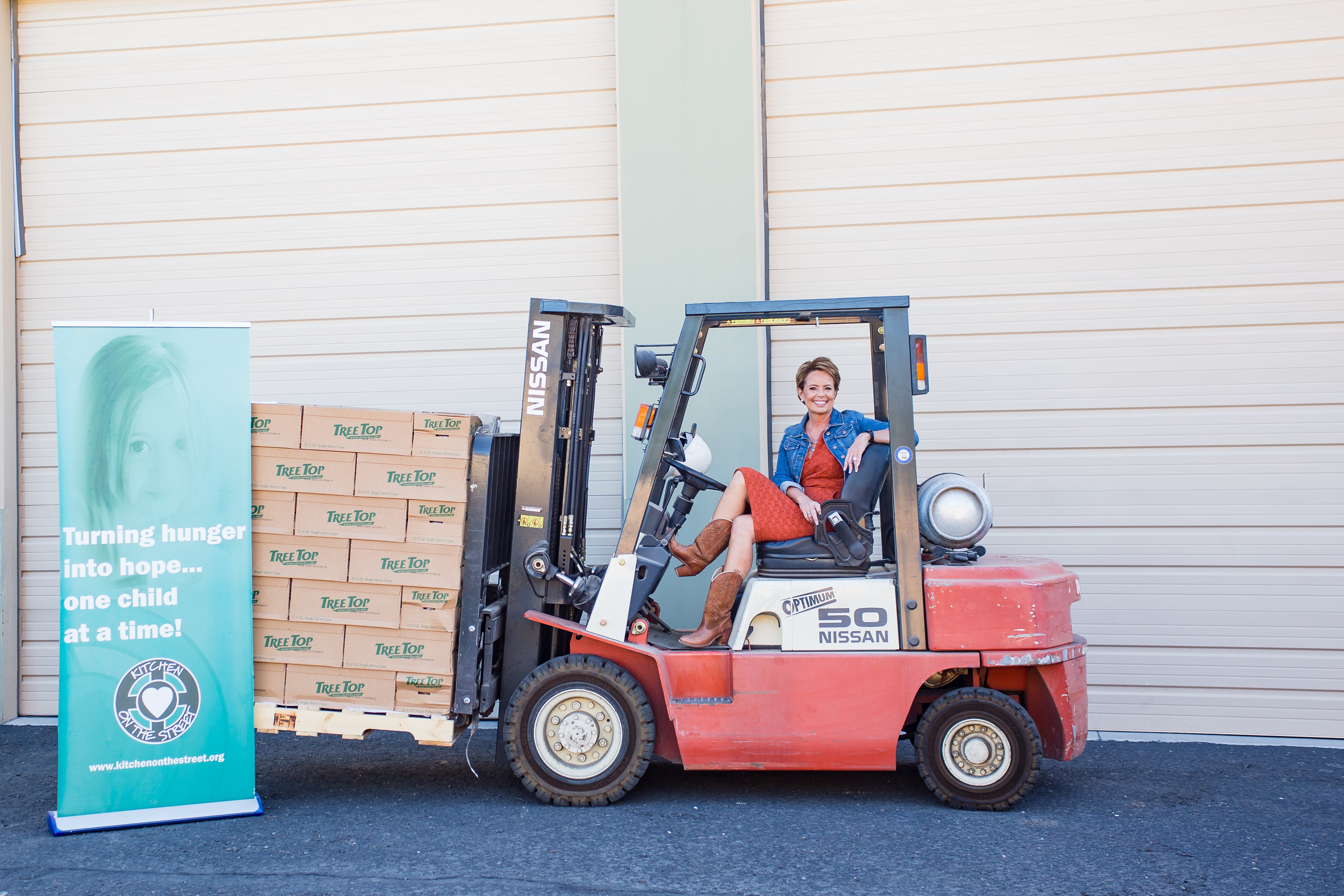 Person operating a forklift with boxes in a warehouse, next to a banner about child hunger, against a white wall.
